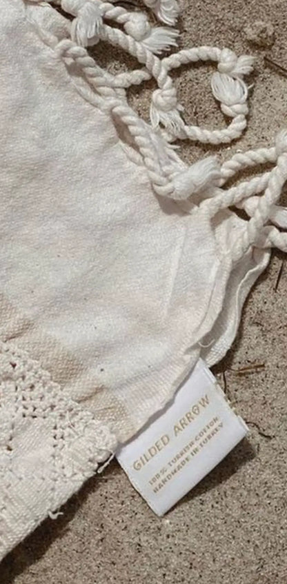 White cotton label with "GILDED ARROW" printed in black, attached to a textured fabric with braided tassels.