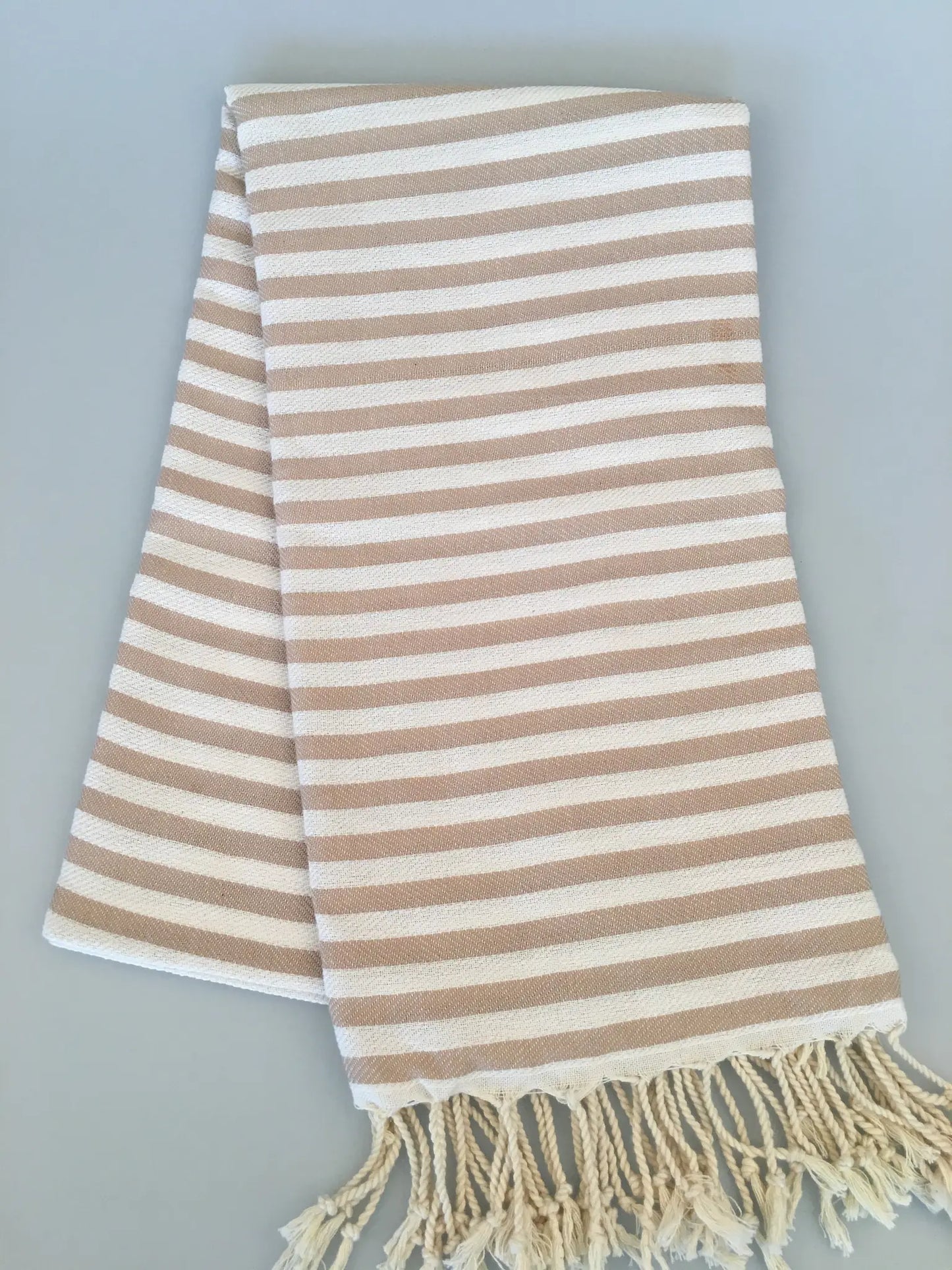 "Beige and cream striped Turkish towel with fringed edges, folded flat on a light gray background."