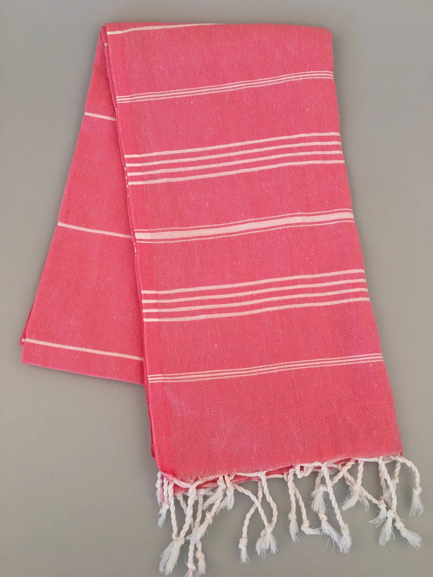 Coral Turkish towel with white stripes and fringed edges, folded on a neutral background.
