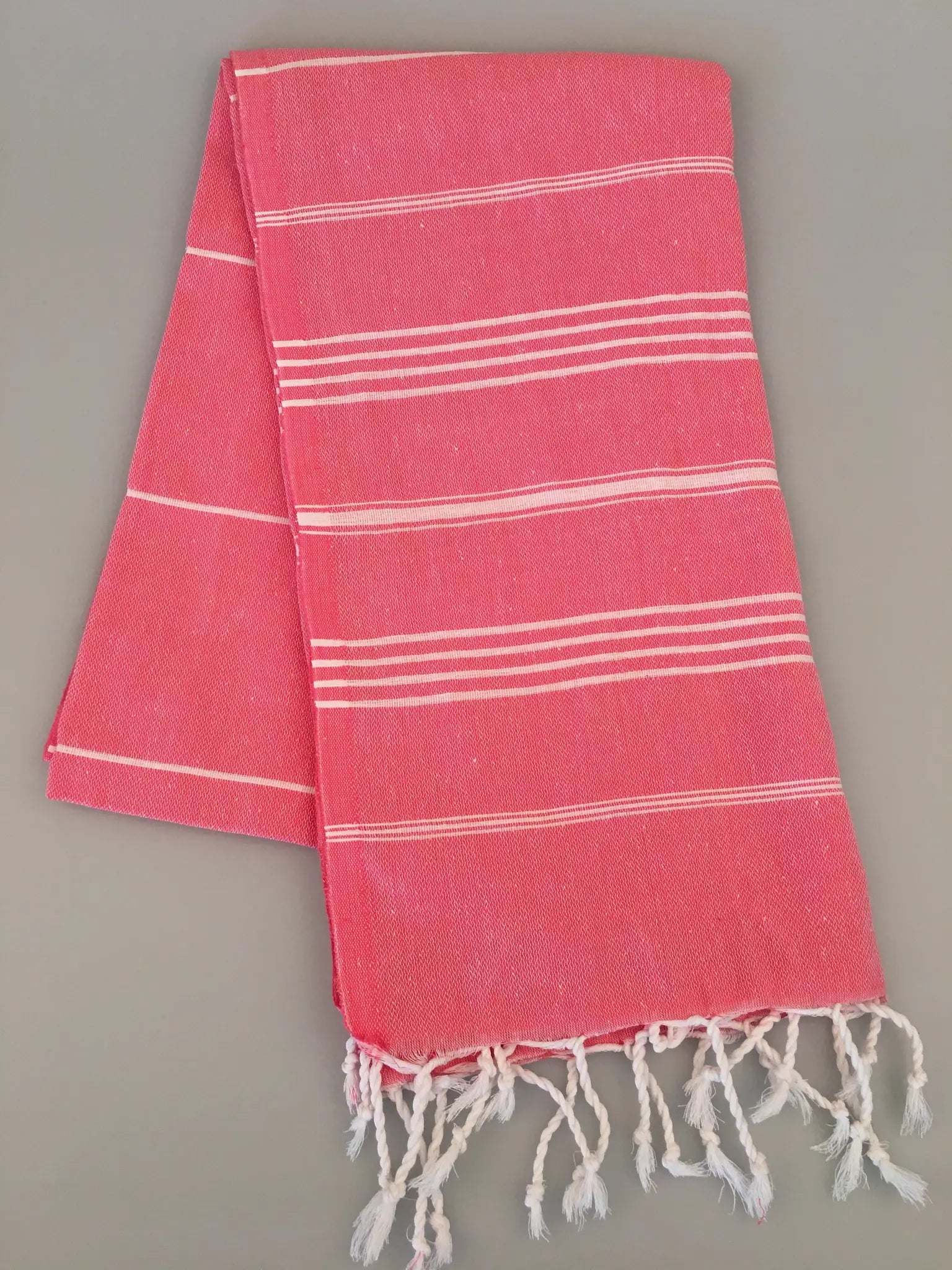 Coral Turkish towel with white stripes and fringed edges, folded on a neutral background.