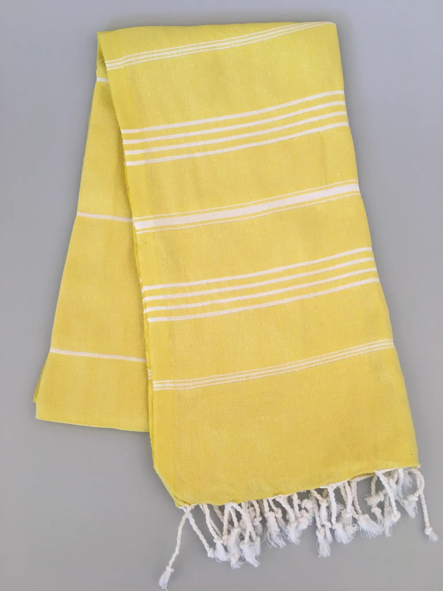 "Yellow Turkish towel with white stripes and fringed edges, laid flat on a gray background."