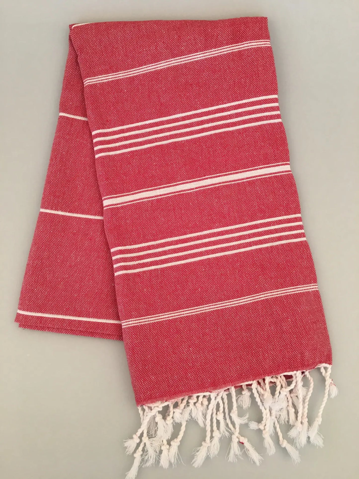 Red Turkish towel with white stripes and fringed ends, folded neatly on a light background.