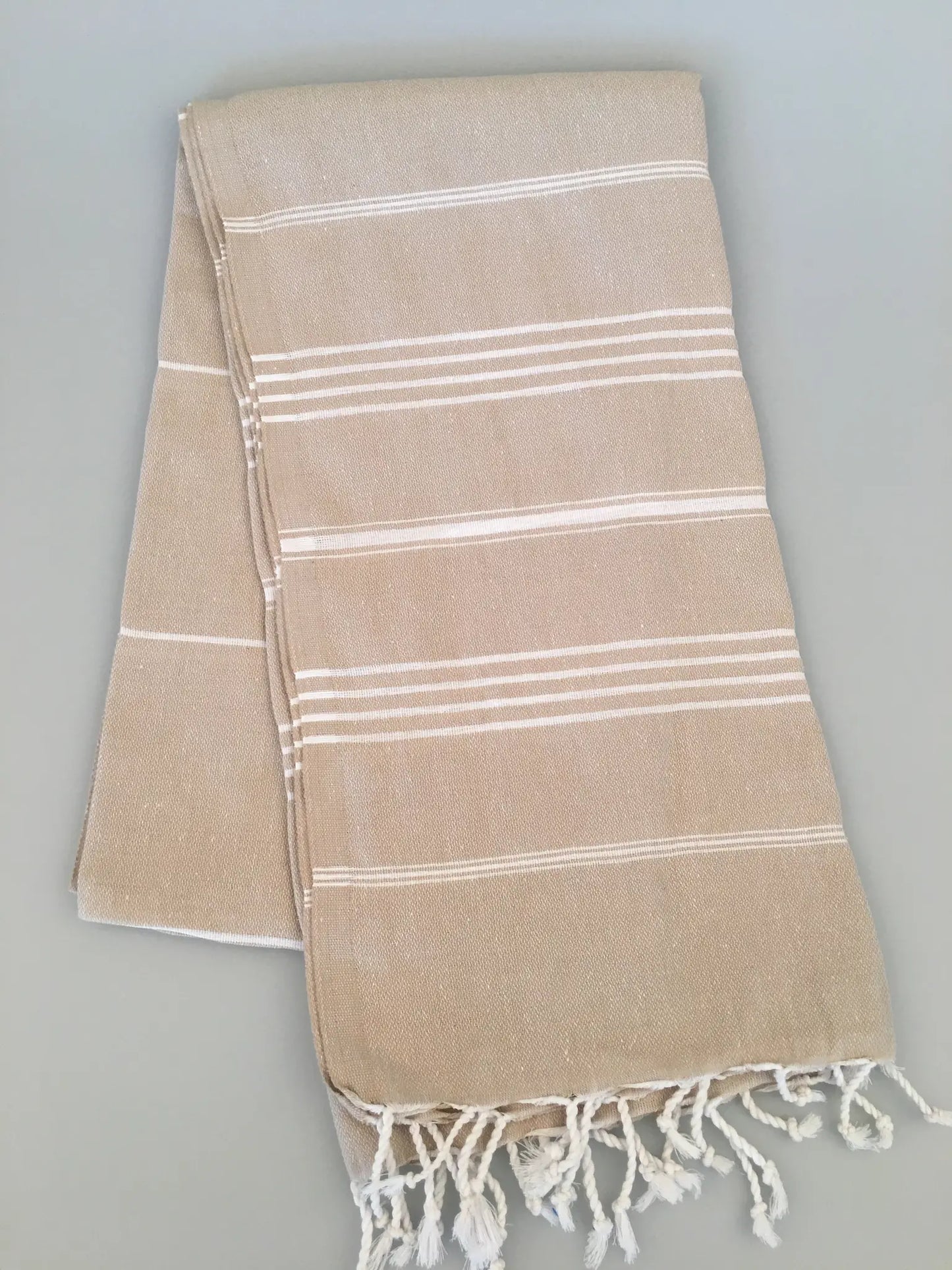 Beige Turkish towel with white stripes and fringed edges, folded neatly on a light gray background.