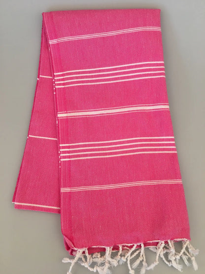 "Pink Turkish towel with white stripes and fringed edges, folded neatly on a gray background."