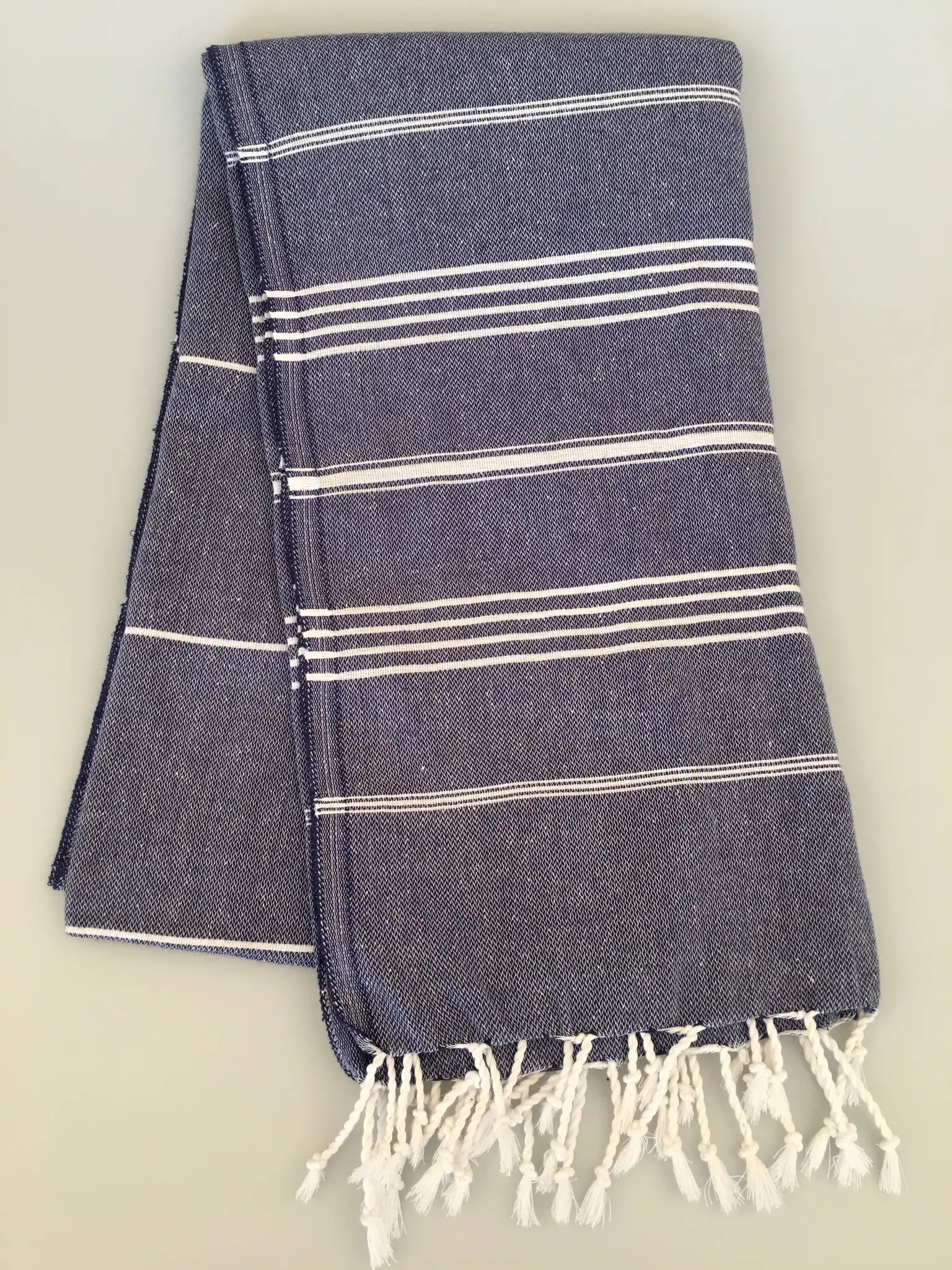 "Dark blue Turkish towel with white stripes and fringed edges, neatly folded on a light background."