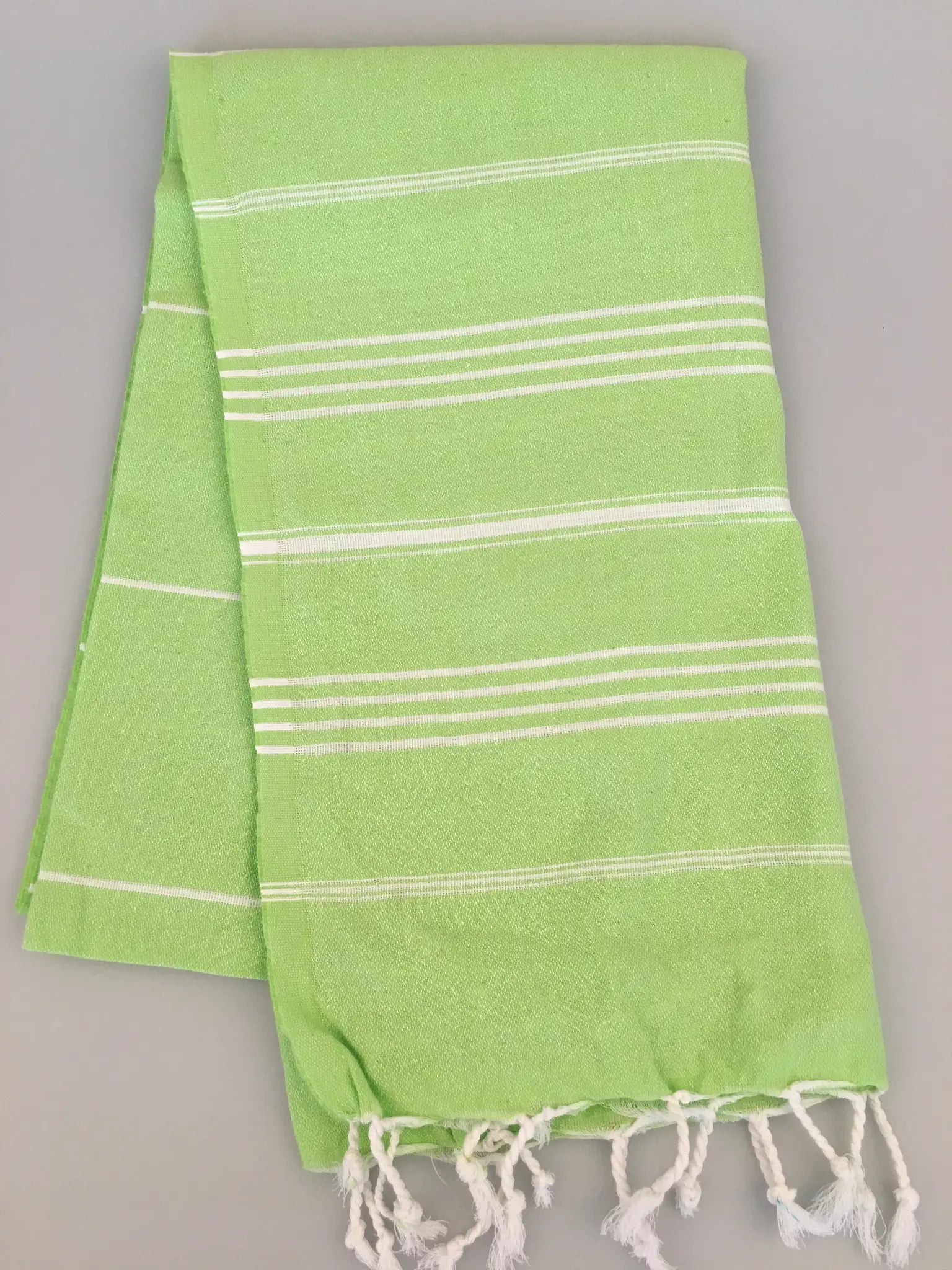 Light green Turkish towel with white stripes and fringed edges, folded on a light gray background.