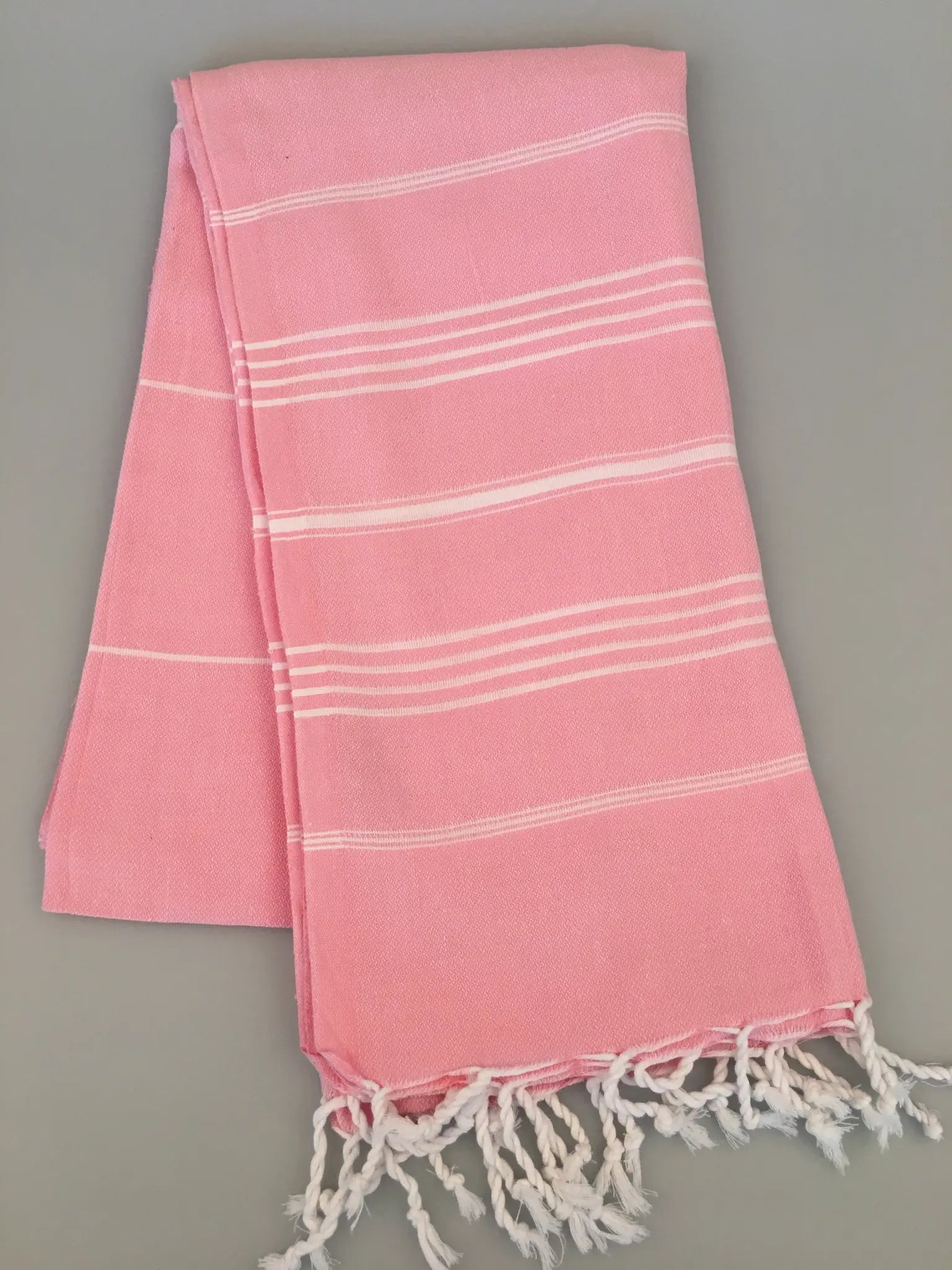 Light pink Turkish towel with white stripes and fringed edges, laid flat on a gray surface.