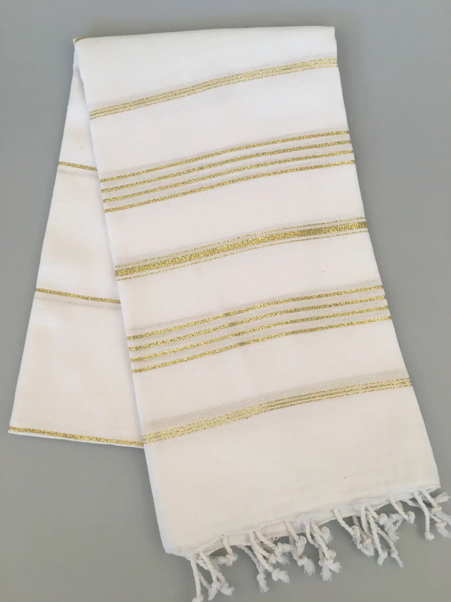 White Turkish towel with gold stripes and fringed edges, folded on a gray background.