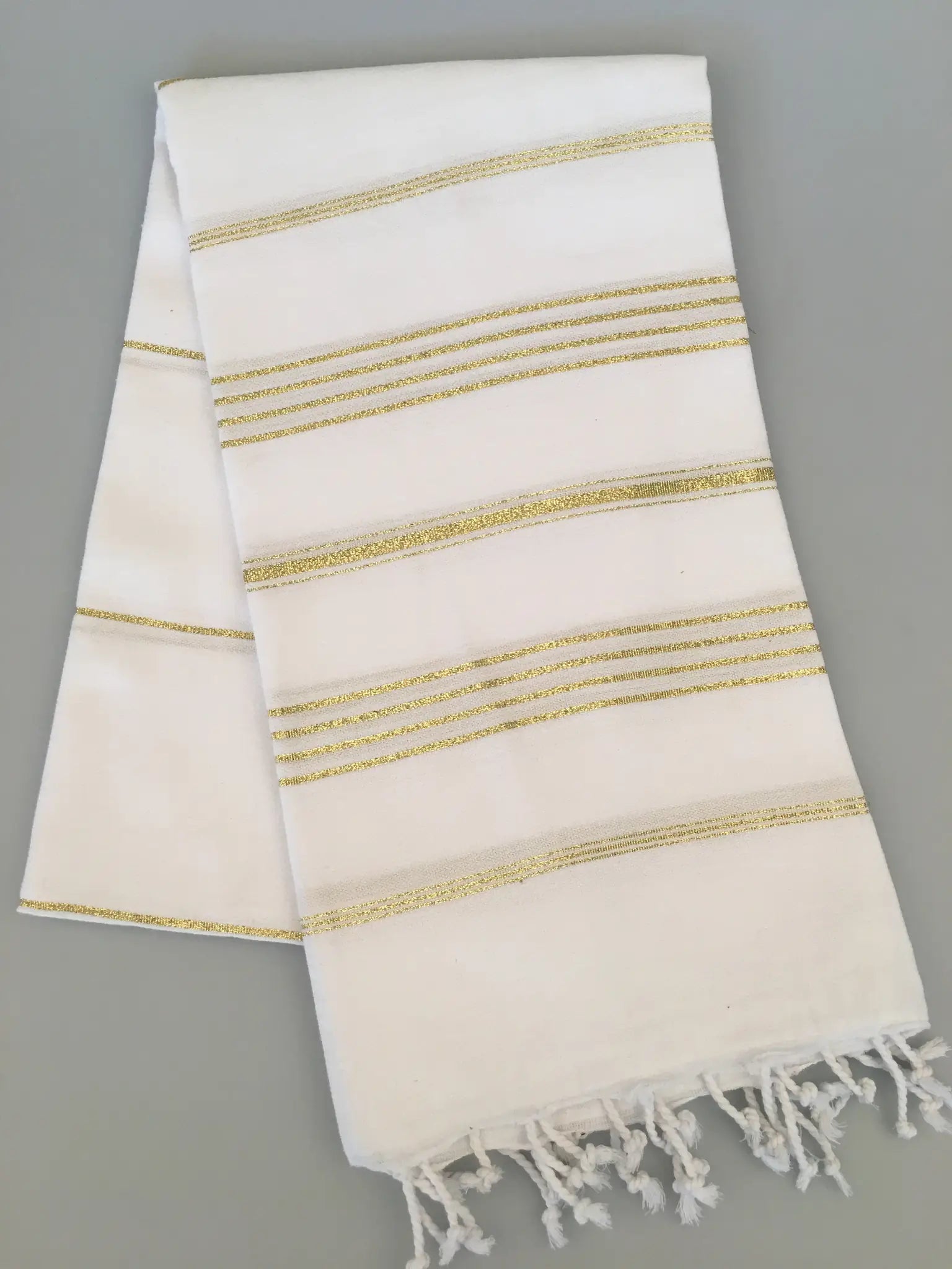 White Turkish towel with gold stripes and fringed edges, folded on a gray background.