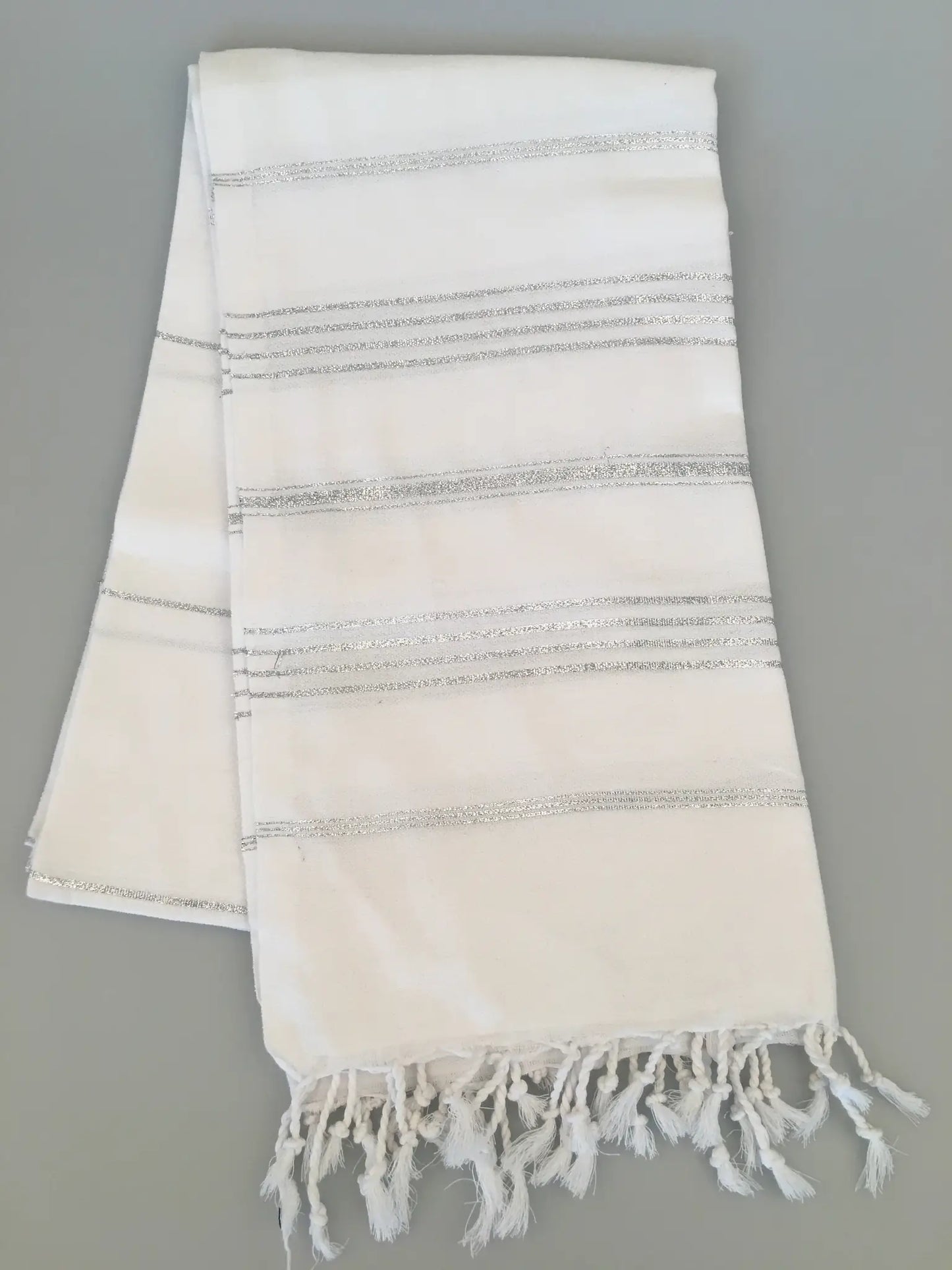 White Turkish towel with silver striped patterns and fringed edges, laid flat on a light gray background.