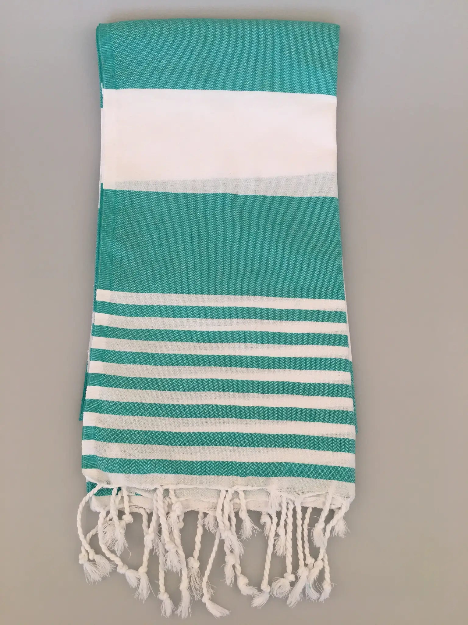 "Turquoise and white striped Turkish towel with fringed edges, folded view on a light background."
