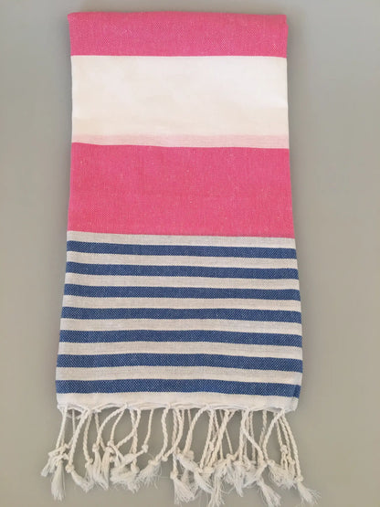 "Tralles Turkish Towel Peshtemal with pink, white, and blue striped pattern, fringed ends, laid flat on a light surface."