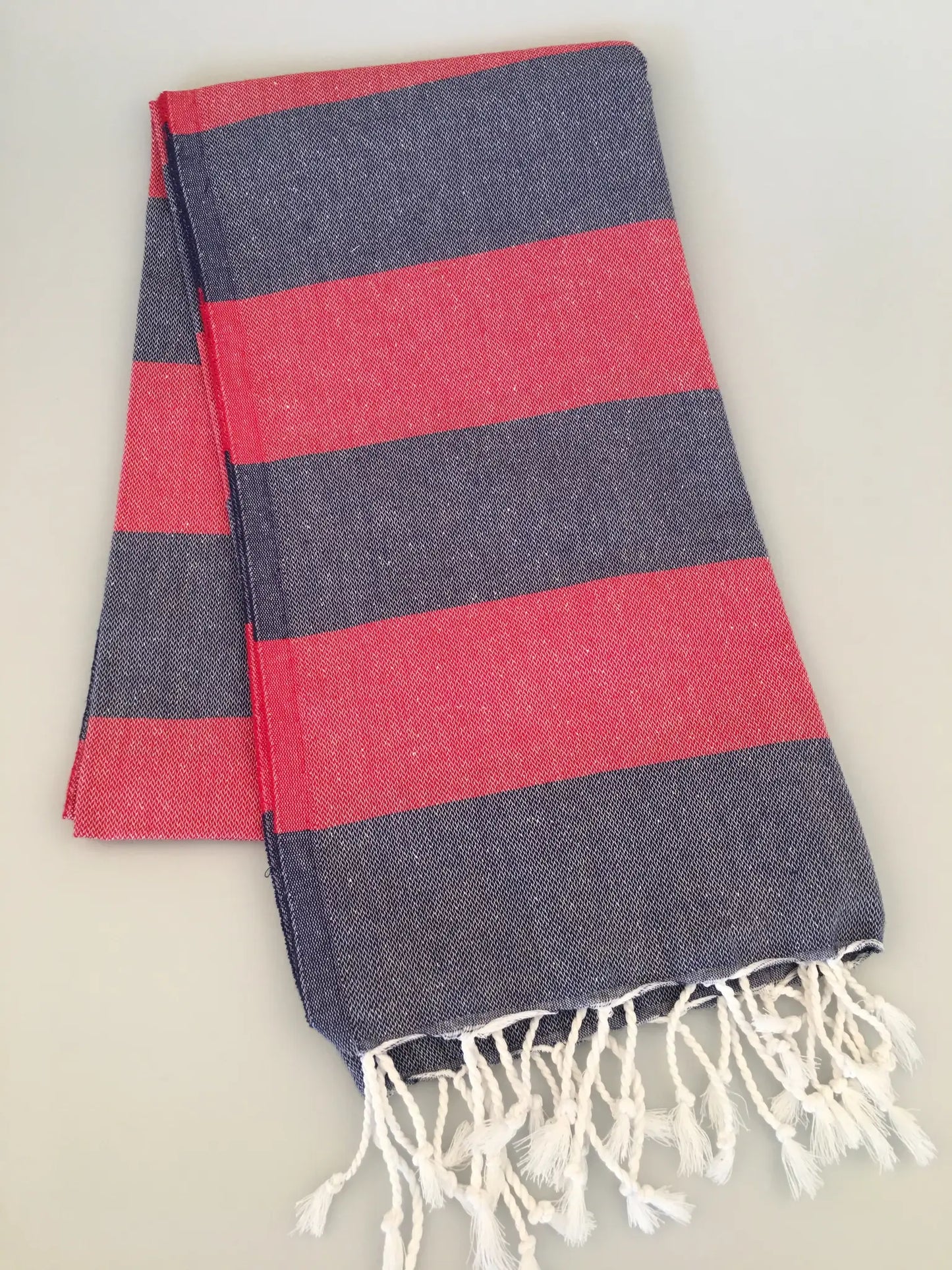 "Pinara Turkish towel peshtamal in navy blue with red stripes and white fringes, folded on a light surface."