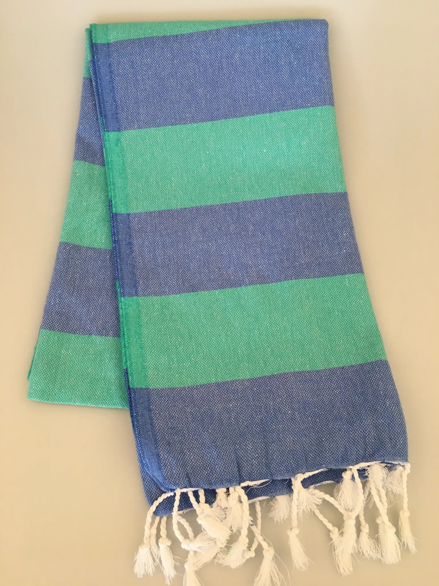 "Blue and green striped Turkish towel with fringed edges, folded neatly on a light background."