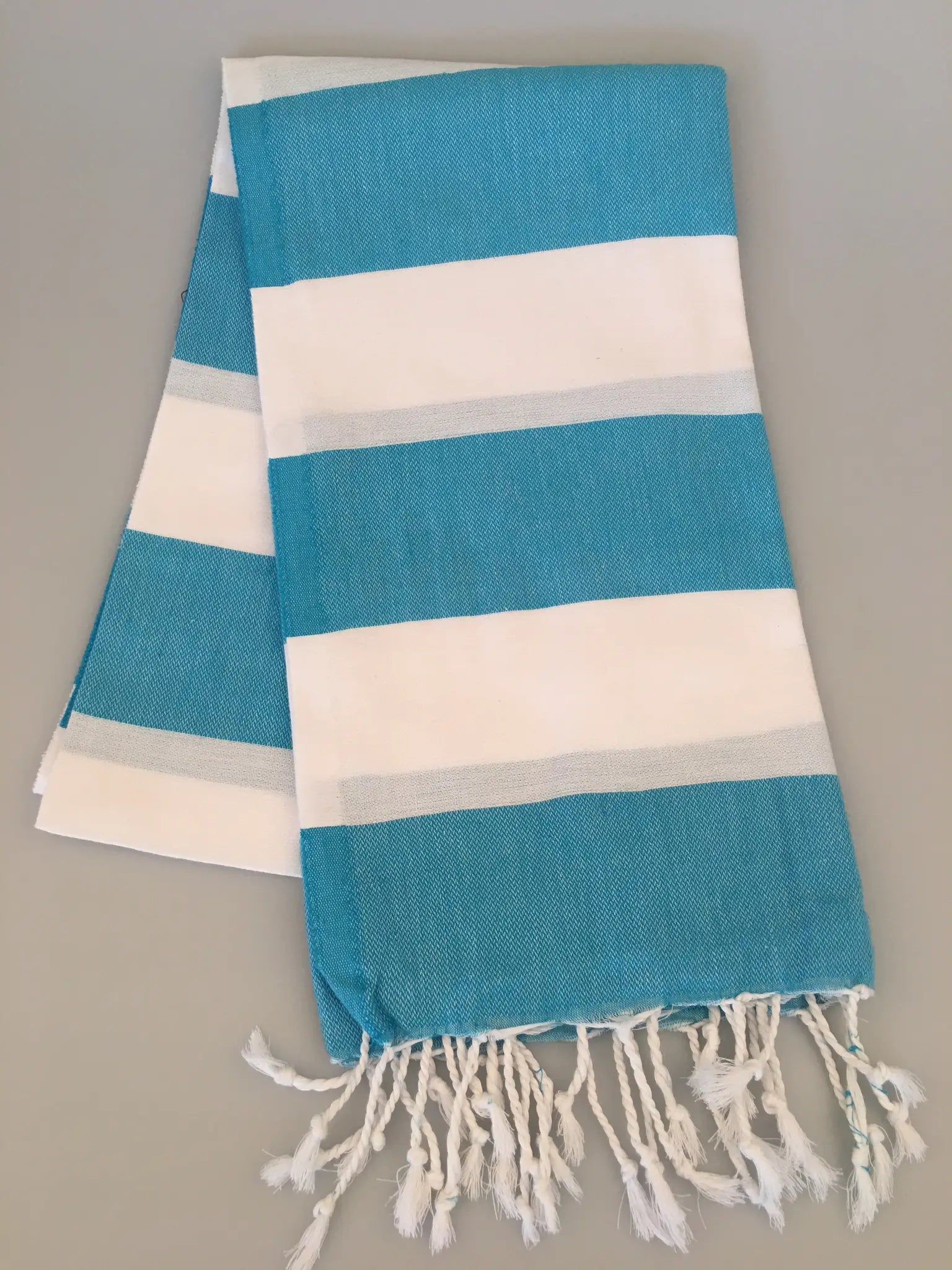 Turquoise and white striped Turkish towel with fringed edges, folded on a light gray background.