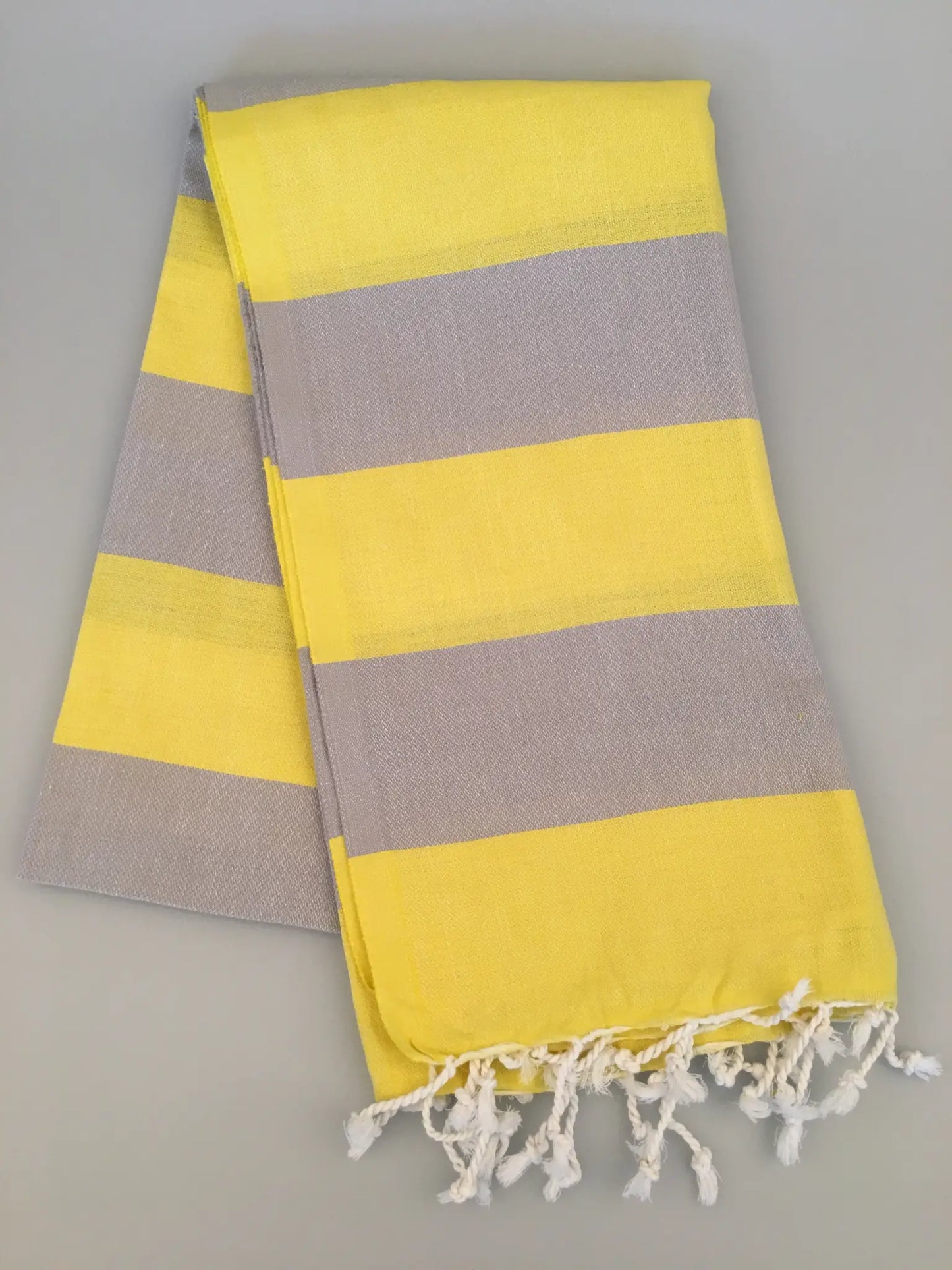"Yellow and gray striped Turkish towel with fringed edges, folded on a light gray background."