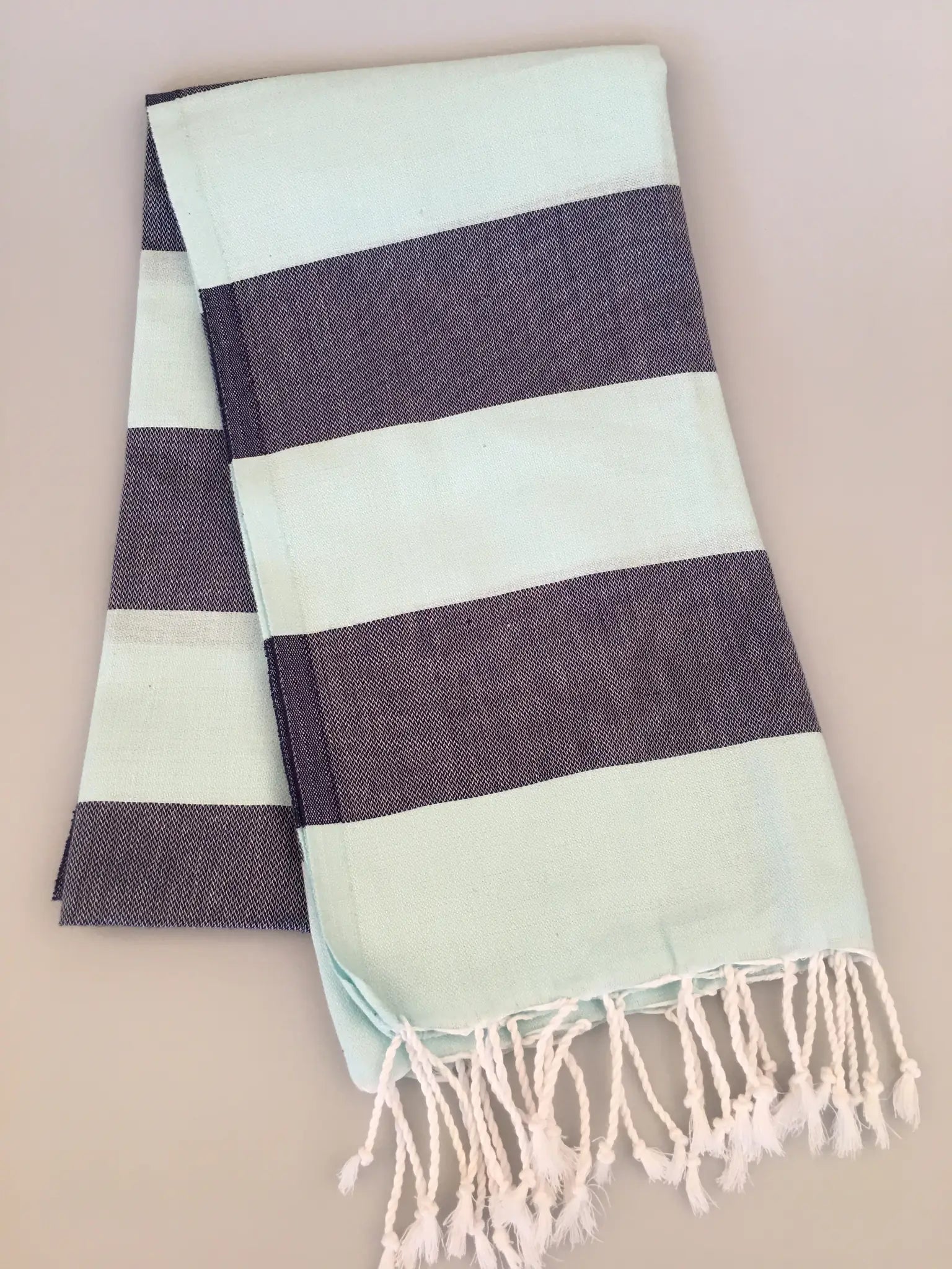 Light turquoise Turkish towel with dark navy stripes and white fringes, folded neatly on a light gray surface.