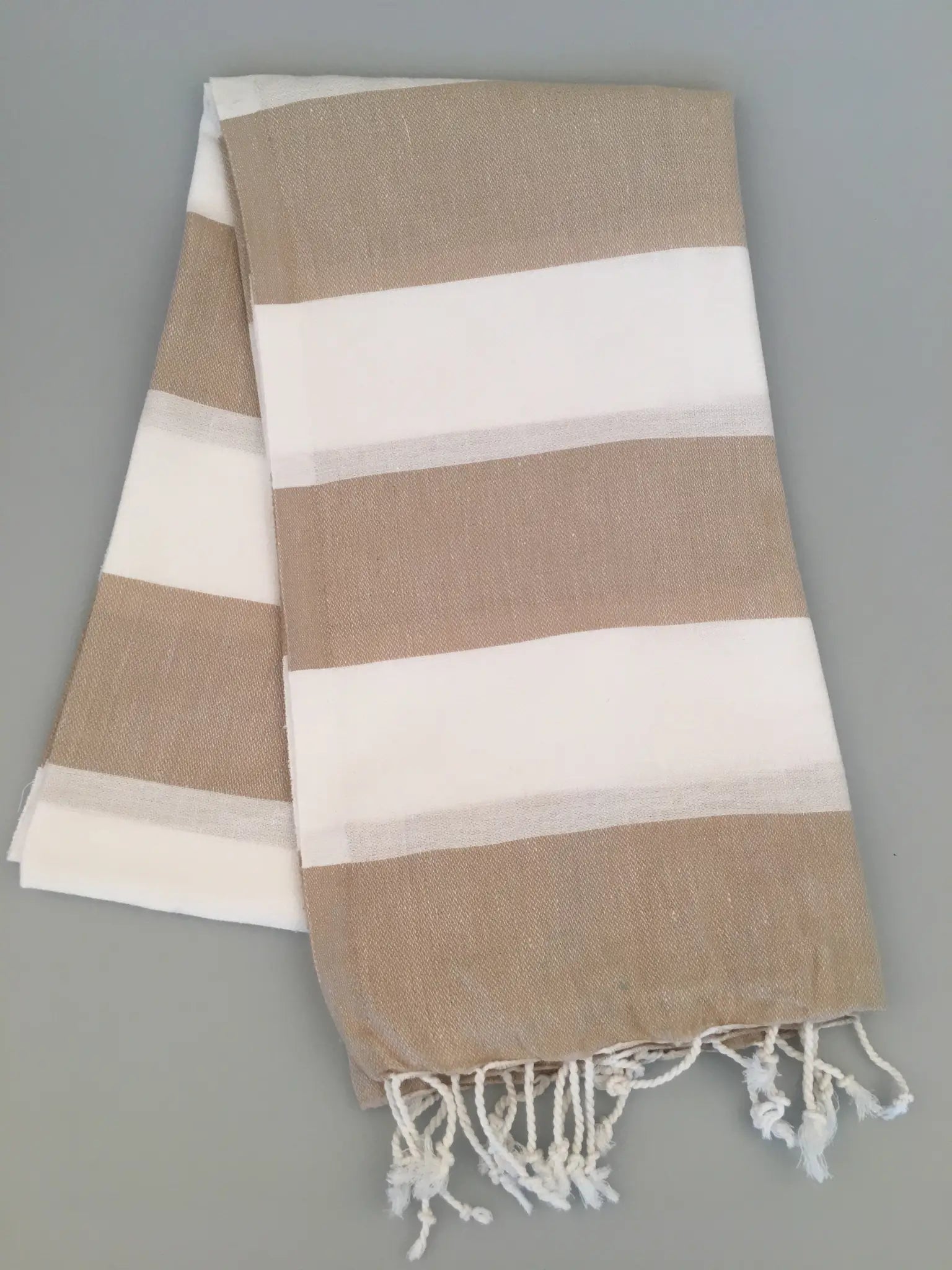 "Pinara Turkish Towel Peshtamal in beige and white stripes, folded with fringed edges, on a light gray background."