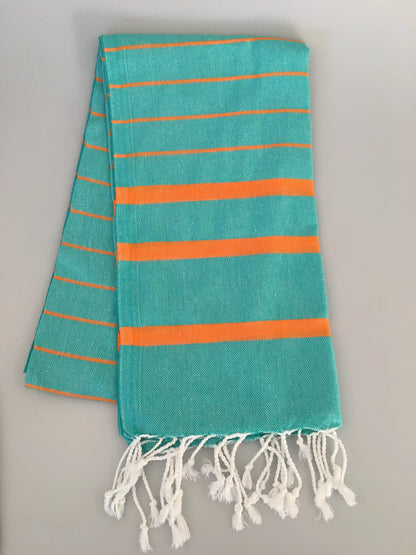Turquoise Turkish towel peshtemal with orange stripes and white fringes, laid flat on a light gray surface.