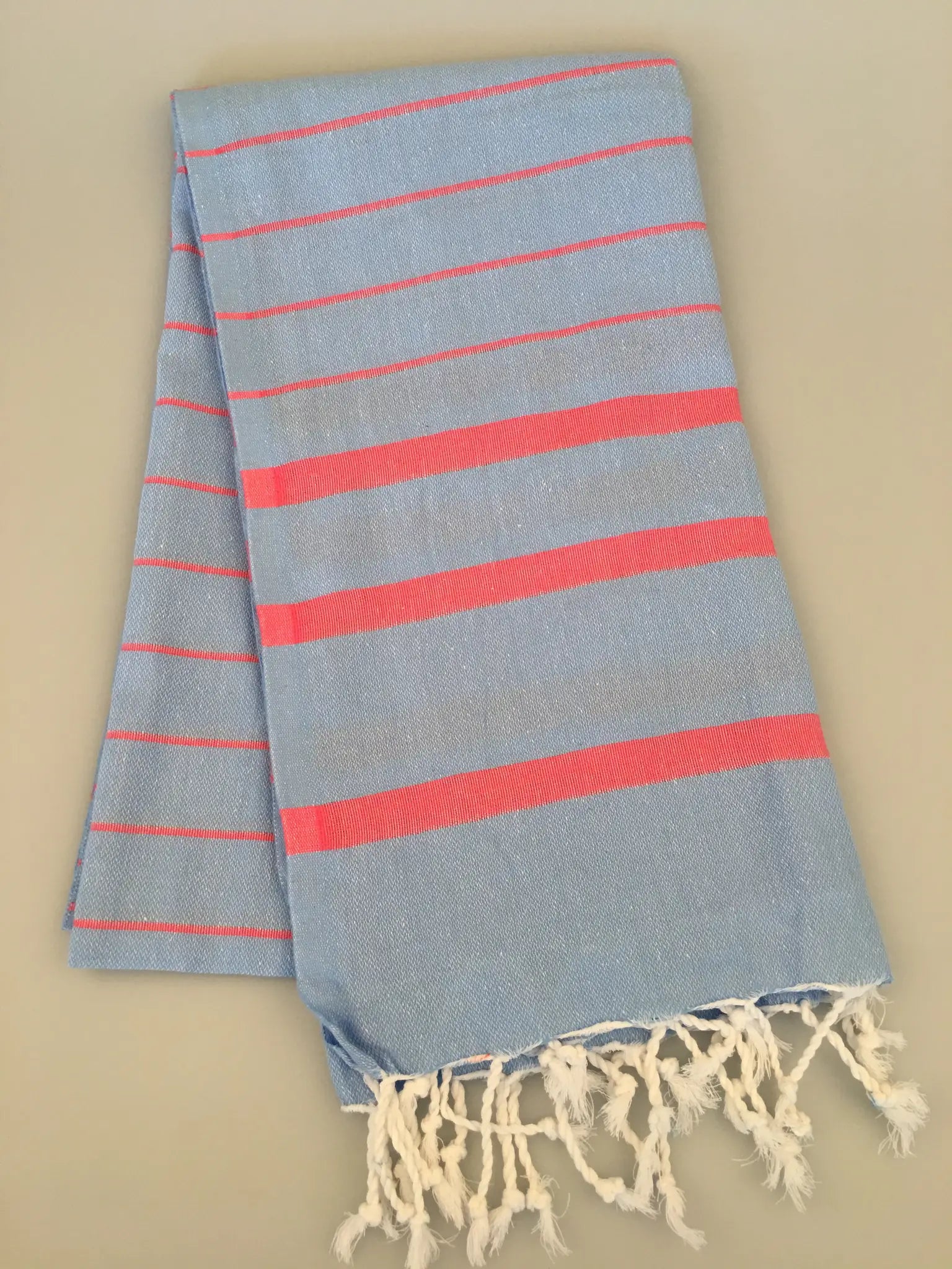 Light blue Turkish towel with pink stripes and fringed edges, laid flat on a light beige background.