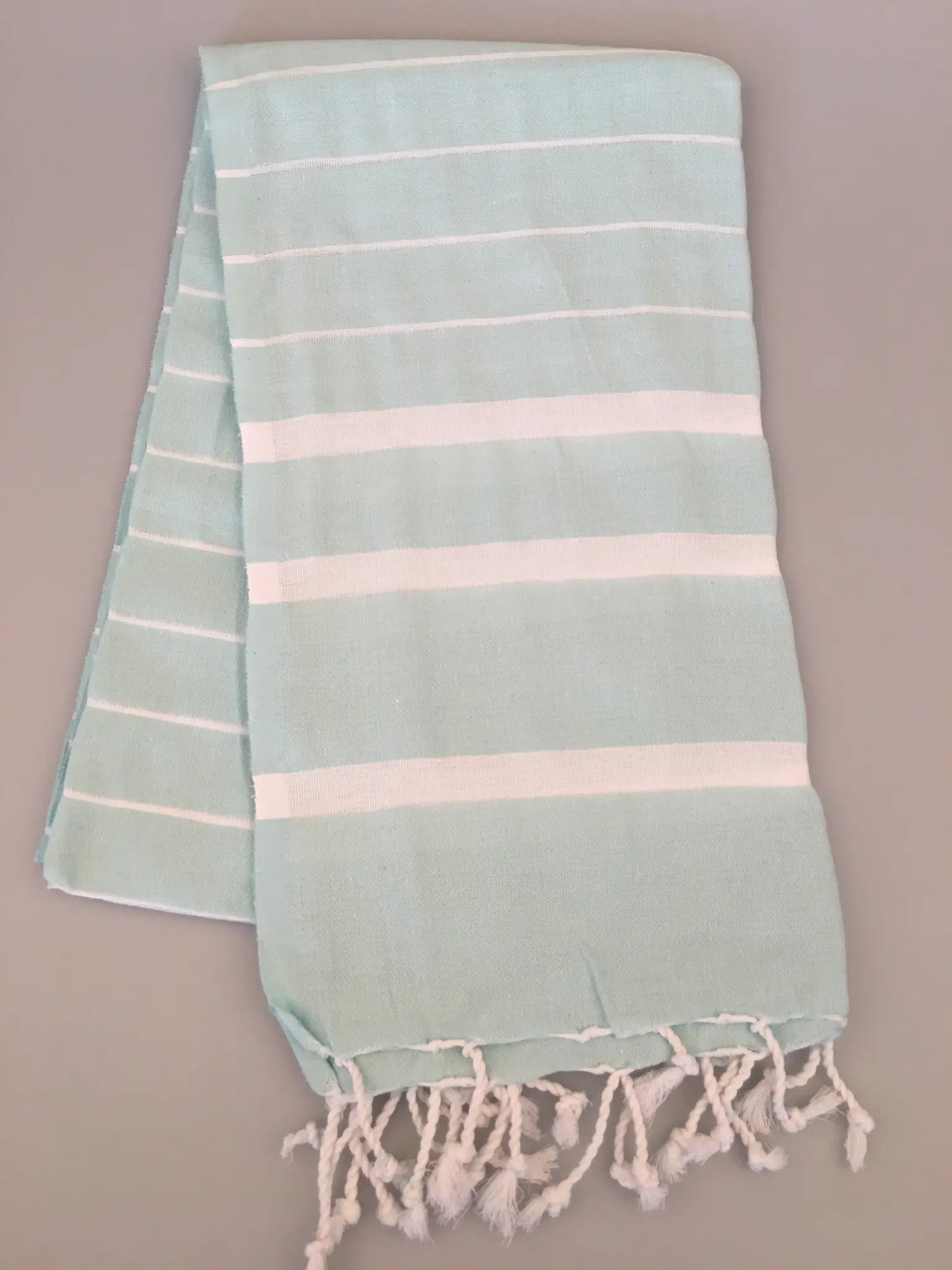 Light blue Turkish towel peshtemal with white stripes and fringed edges, displayed flat on a gray background.