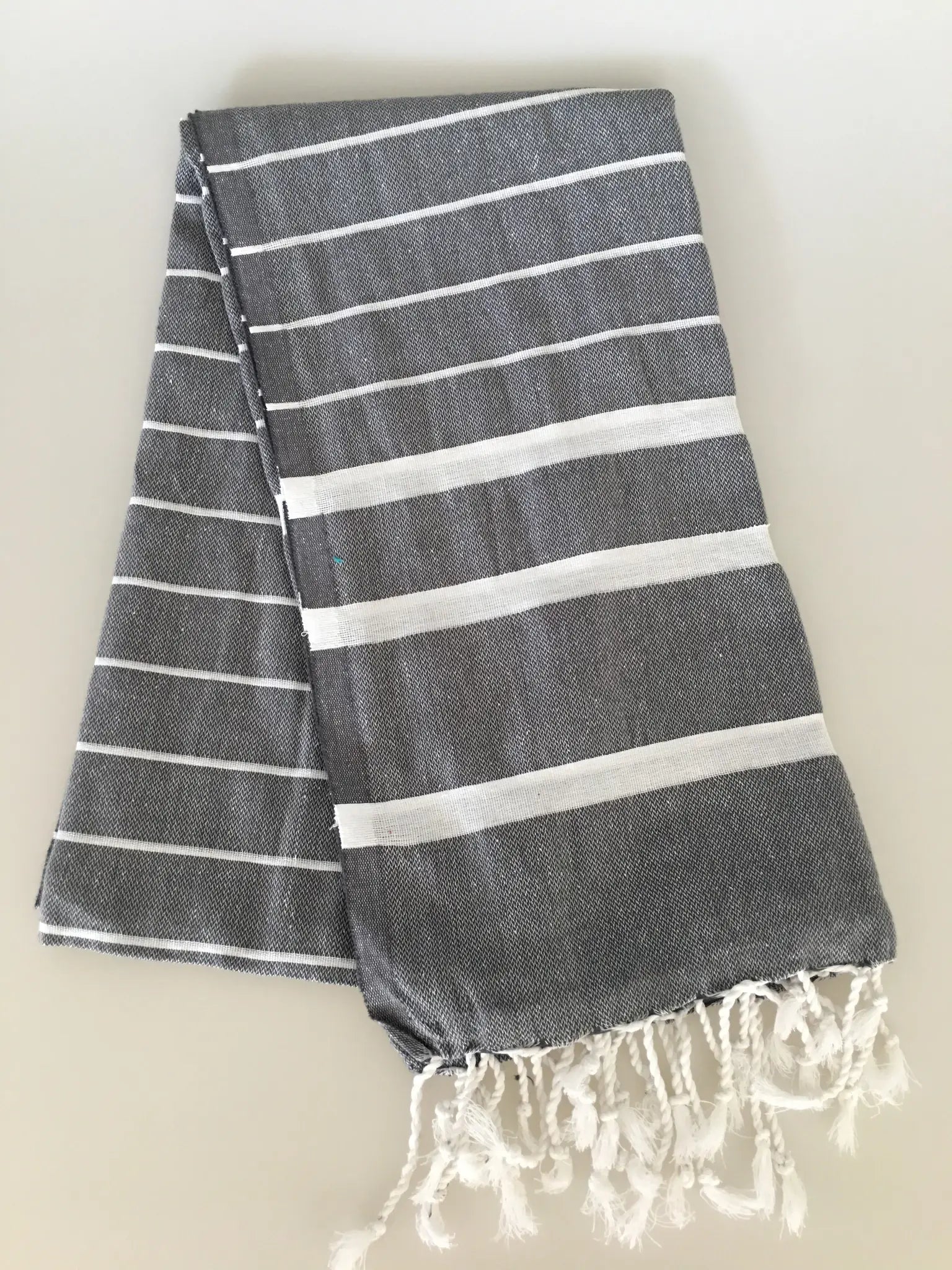 "Gray Turkish towel peshtemal with white stripes and fringed edges, neatly folded on a light background."