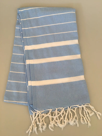 Light blue Turkish towel peshtemal with white stripes, featuring fringed edges, laid out flat on a neutral background.