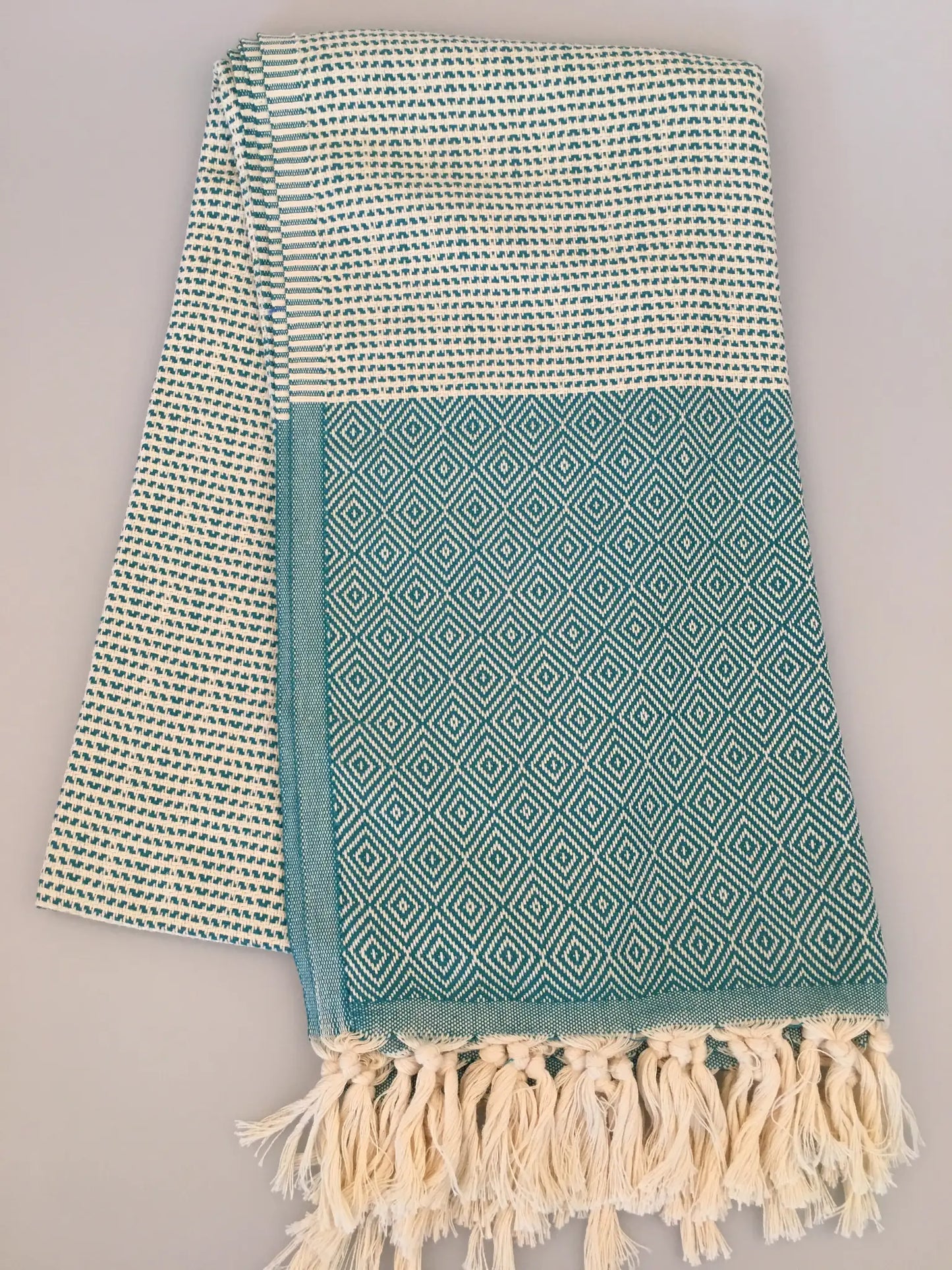 "Turquoise and cream Turkish towel with diamond pattern and fringed edges, folded on a light background."