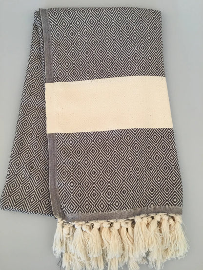 "Gray Turkish towel with diamond pattern, cream stripe, and fringed edges, folded on a light background."
