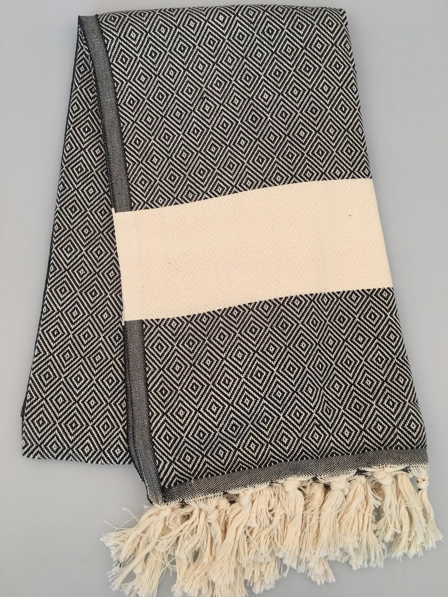"Black and white diamond-patterned Turkish towel with cream band and fringed edges, folded on a gray background."