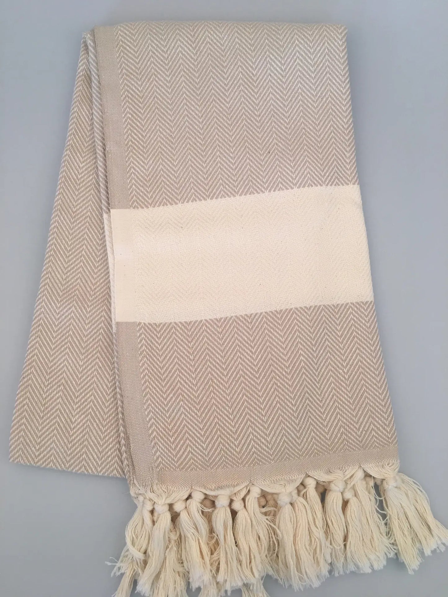 Beige Turkish towel peshtemal with herringbone pattern, cream stripe, and fringed edges, laid flat on a gray background.