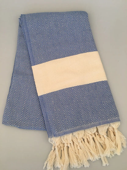 Blue and white chevron-patterned Turkish towel with cream-colored band and fringed edges, folded on a light background.