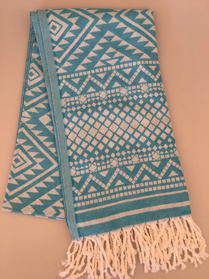 Turquoise jacquard peshtemal towel with geometric patterns, featuring white fringes at one end, laid flat.