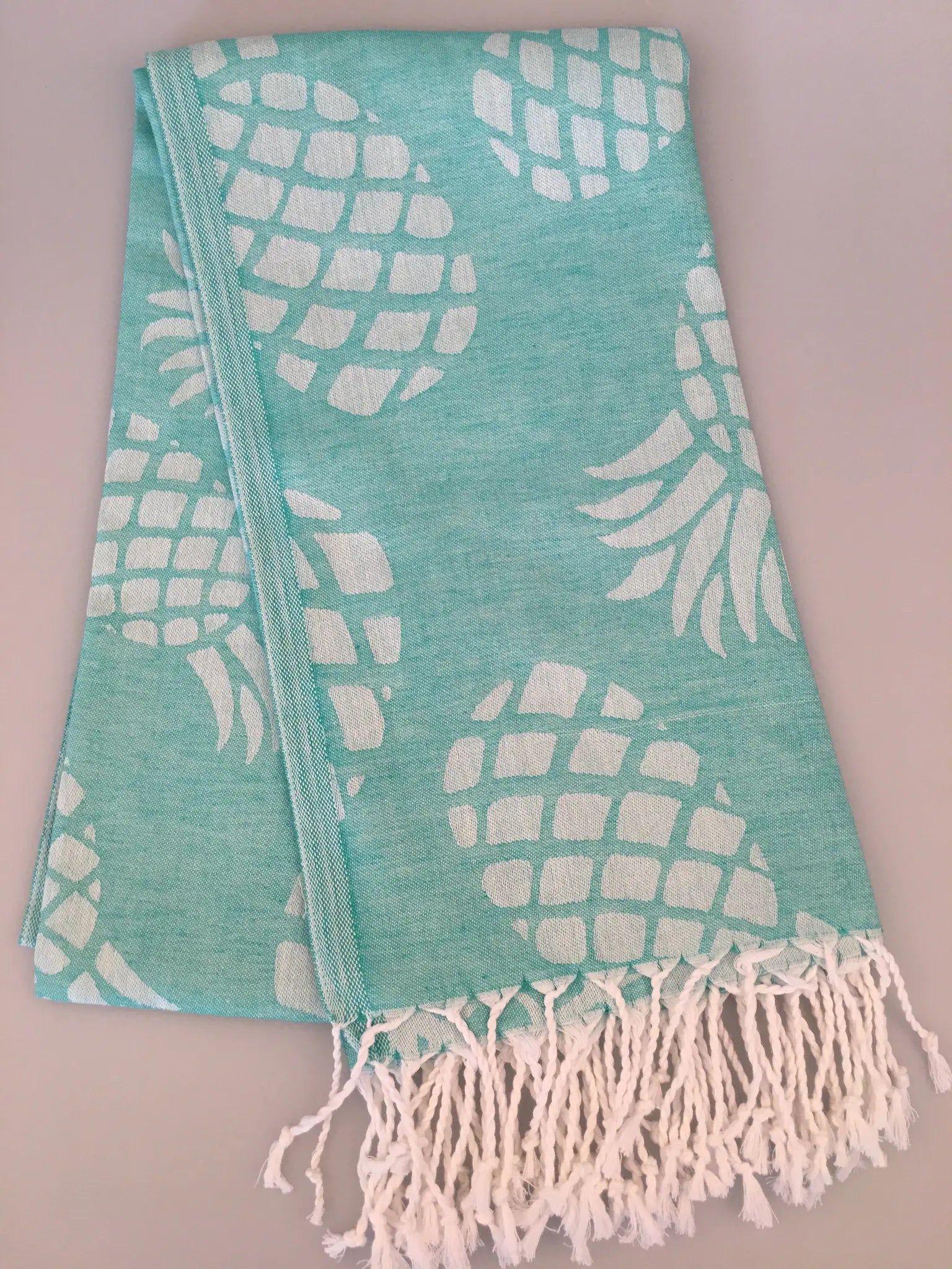 Turquoise Turkish towel with white jacquard pineapple pattern and fringed edges, laid flat on a light background.