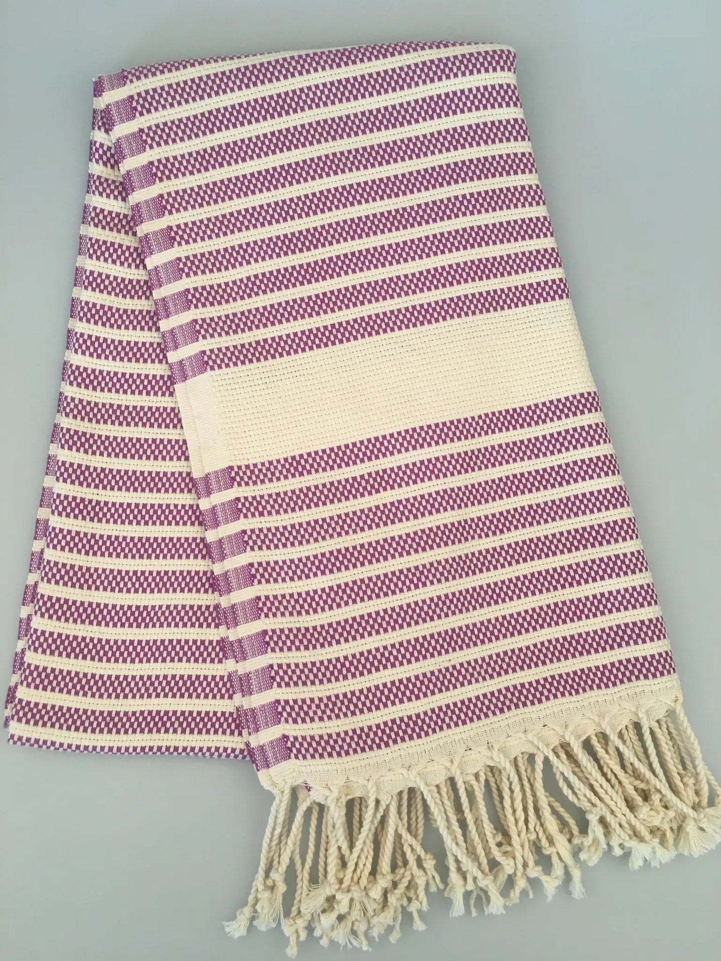 Purple and cream striped Turkish towel with fringed edges, laid flat on a light gray background.