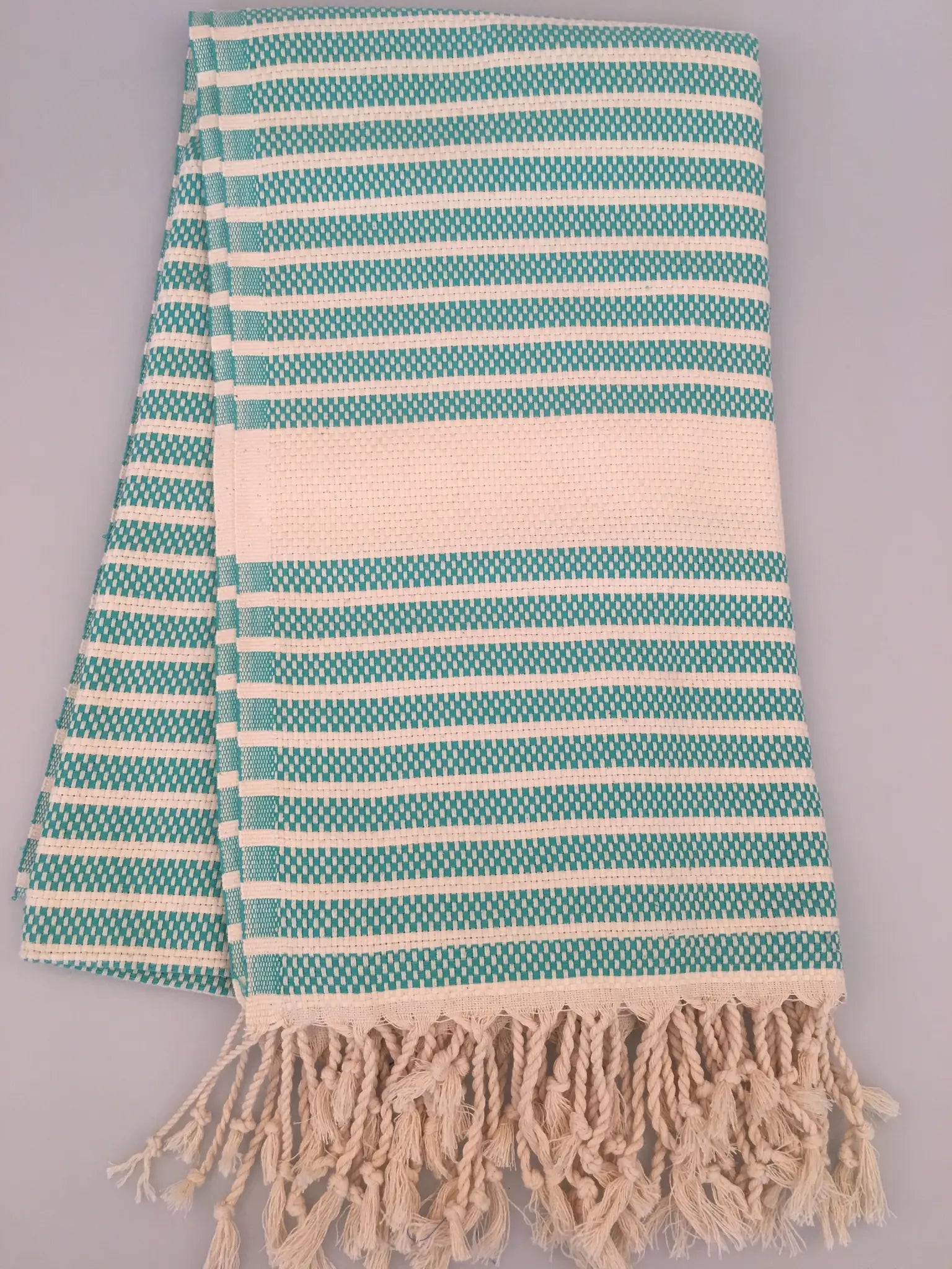 Turquoise and white striped Turkish towel with fringe edges, laid flat on a light background.