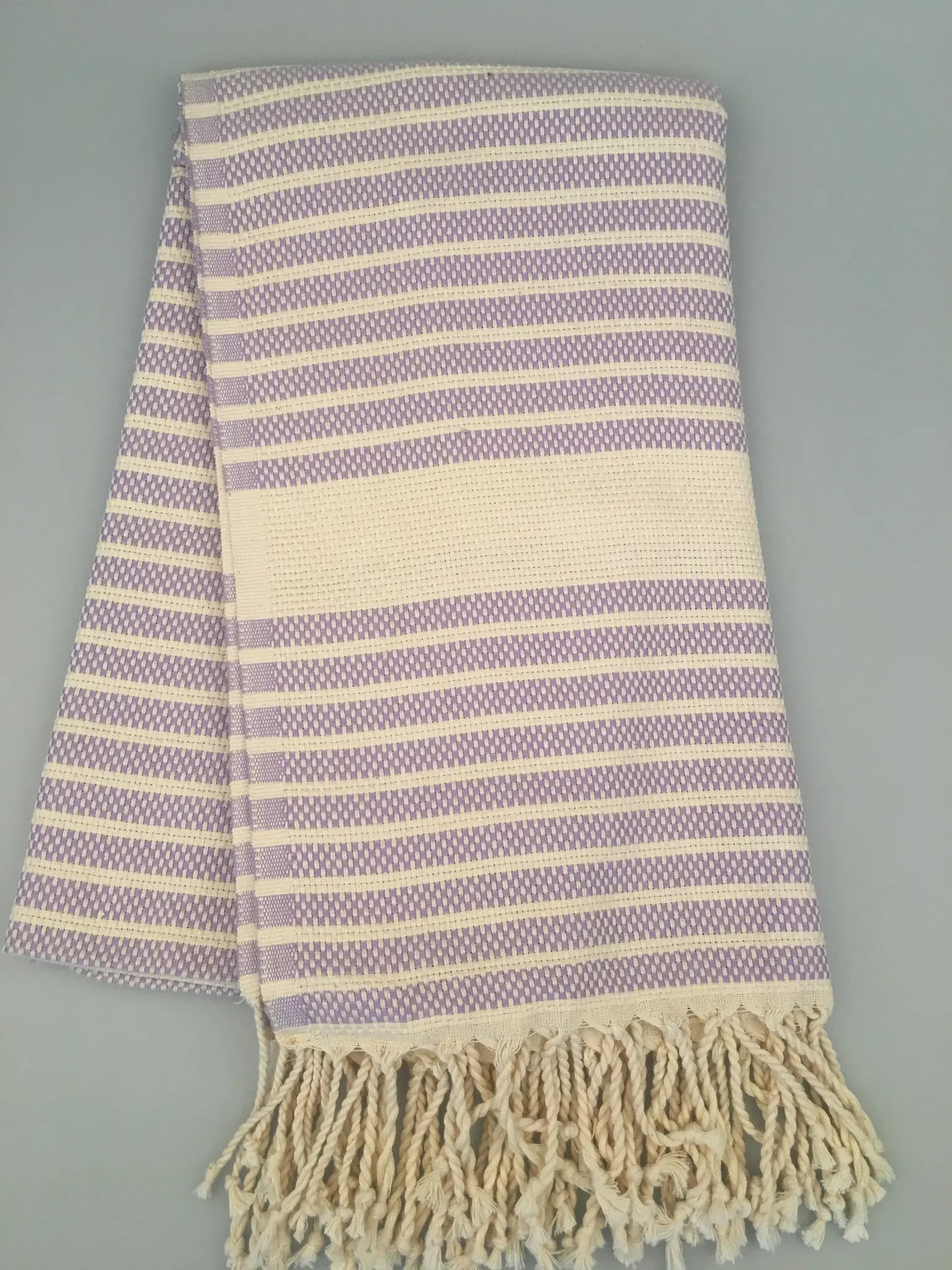 "Light purple and cream striped peshtemal towel with fringed edges, laid flat on a light gray background."