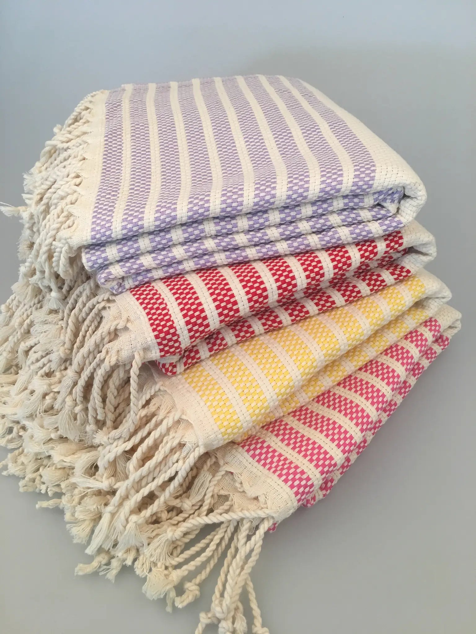 Stack of Hittite Turkish towels in lavender, red, yellow, and pink stripes, with knotted fringe edges on a light background.