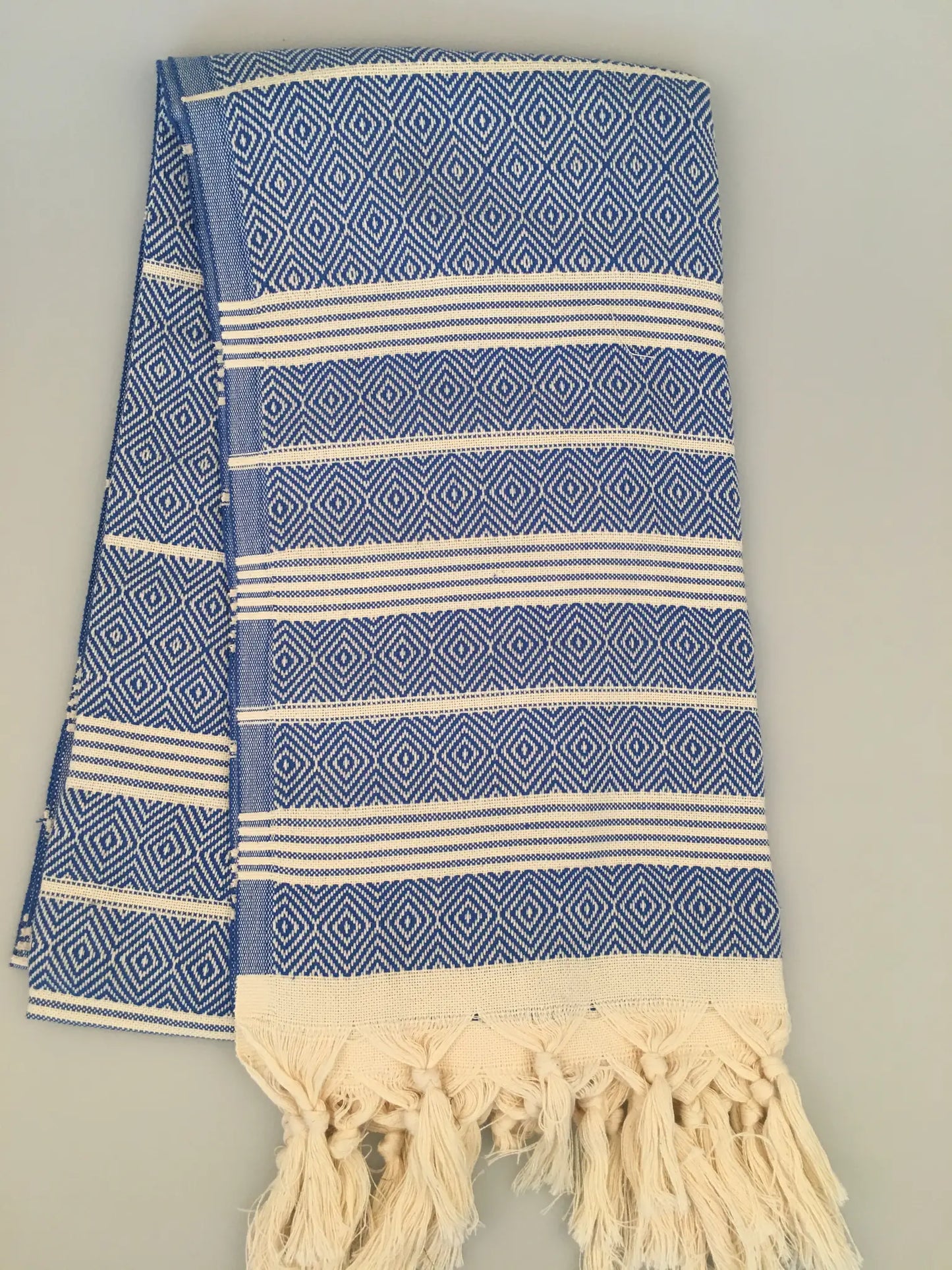 "Lycia Xanthos Turkish towel peshtemal in blue and white diamond pattern with fringed edges, folded on a light surface."