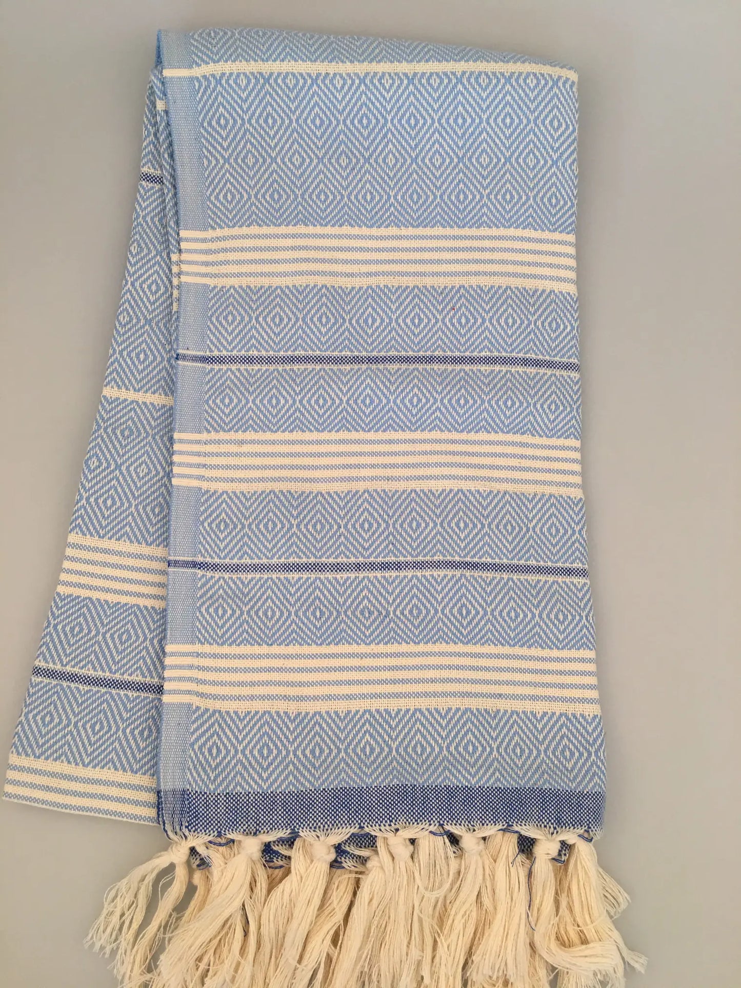 Light blue Turkish towel with white diamond patterns and fringed edges, laid flat on a light gray background.