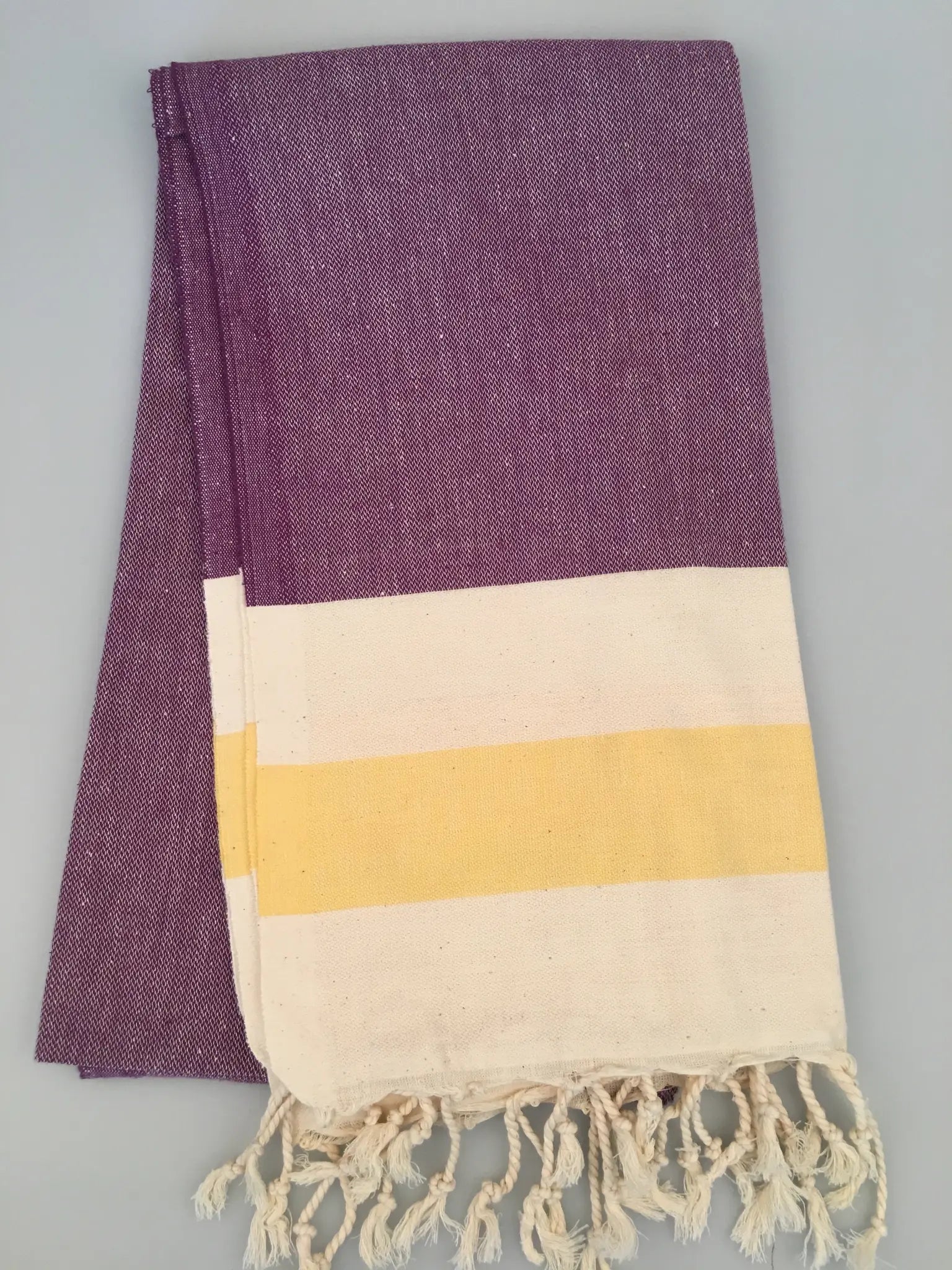 "Purple and cream Turkish towel with yellow stripes and fringed edges, folded on a light gray background."