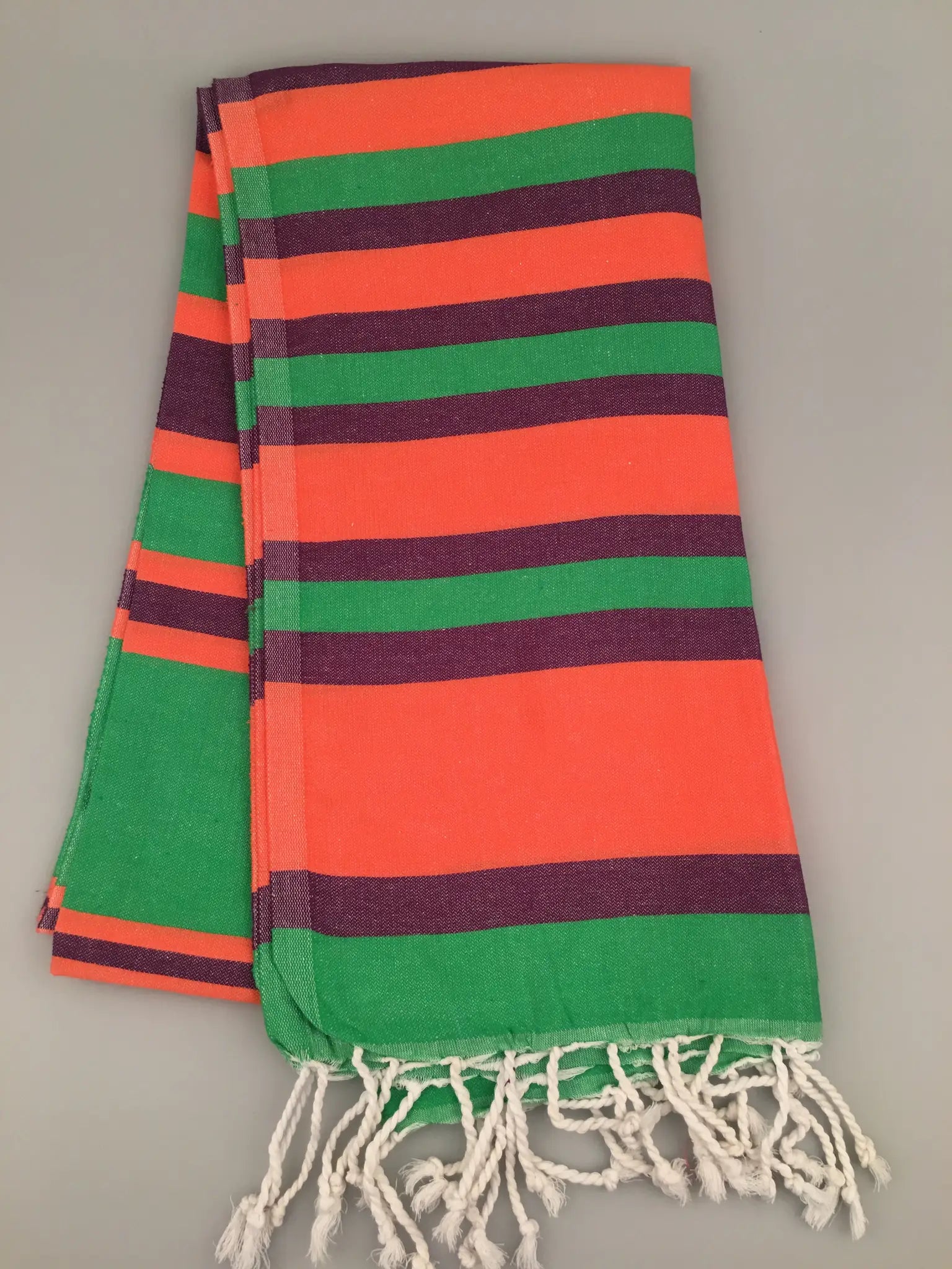 "Striped Aizanoi Turkish towel in orange, green, and purple with knotted fringes, folded flat on a gray surface."