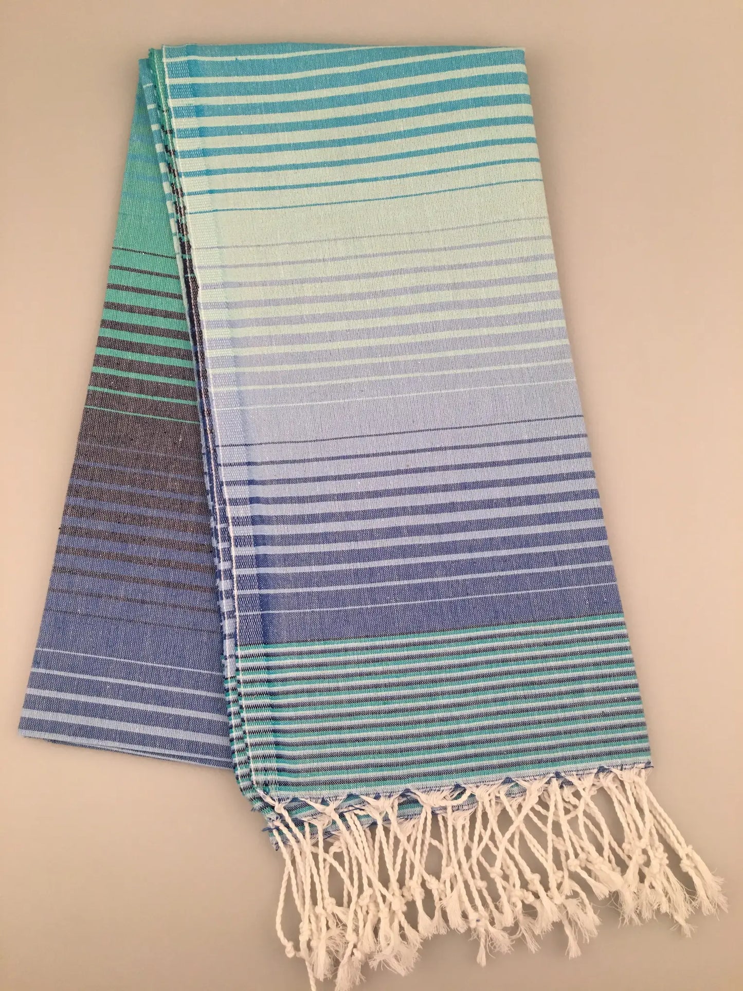 "Striped Turkish towel in gradient shades of blue, featuring white fringes at one end, laid flat on a light background."