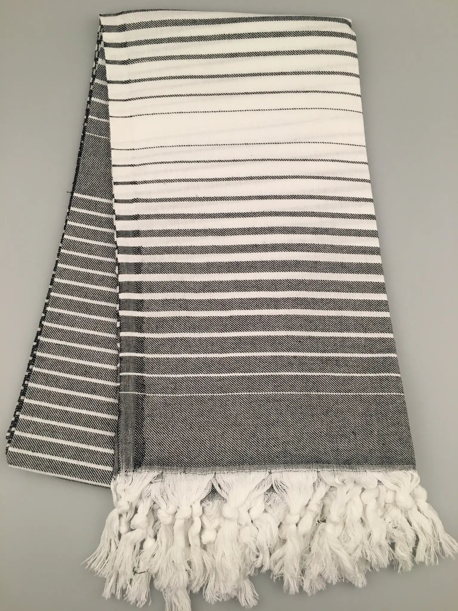"Telmessos Turkish towel peshtemal in gray and white stripes, featuring fringed edges, laid flat on a gray background."