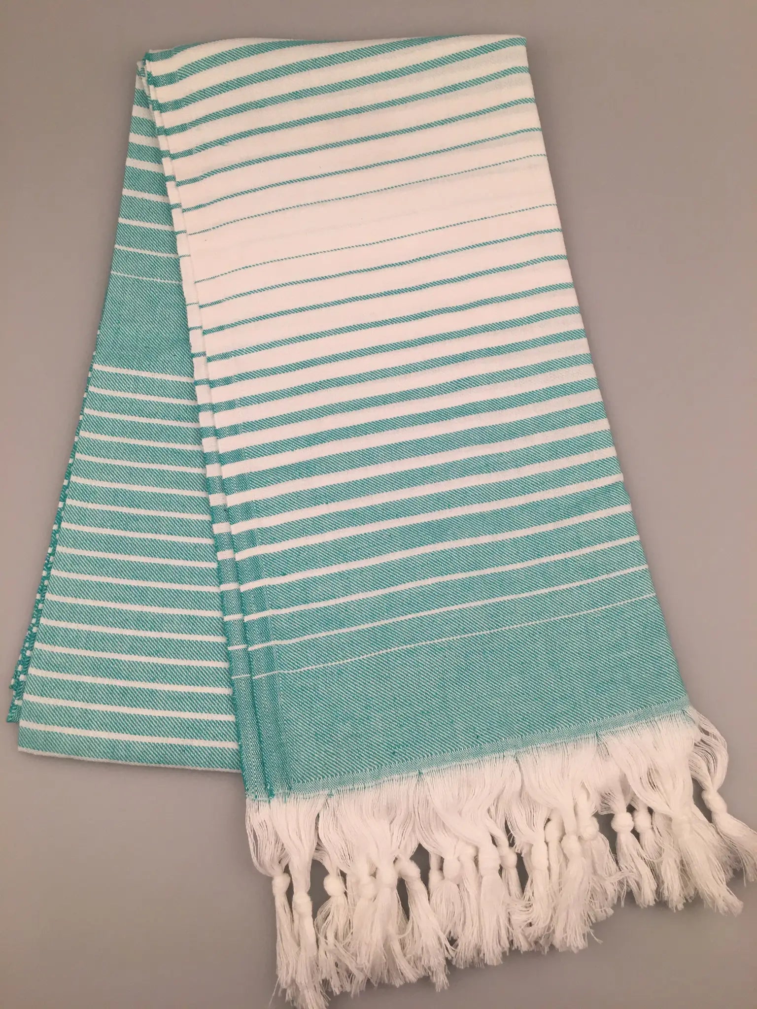 Turquoise and white striped Turkish towel with fringed edges, laid flat on a gray background.