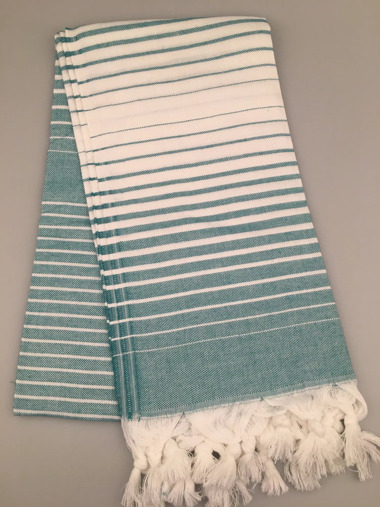 Aqua and white striped Turkish towel with fringed edges, neatly folded on a gray background.