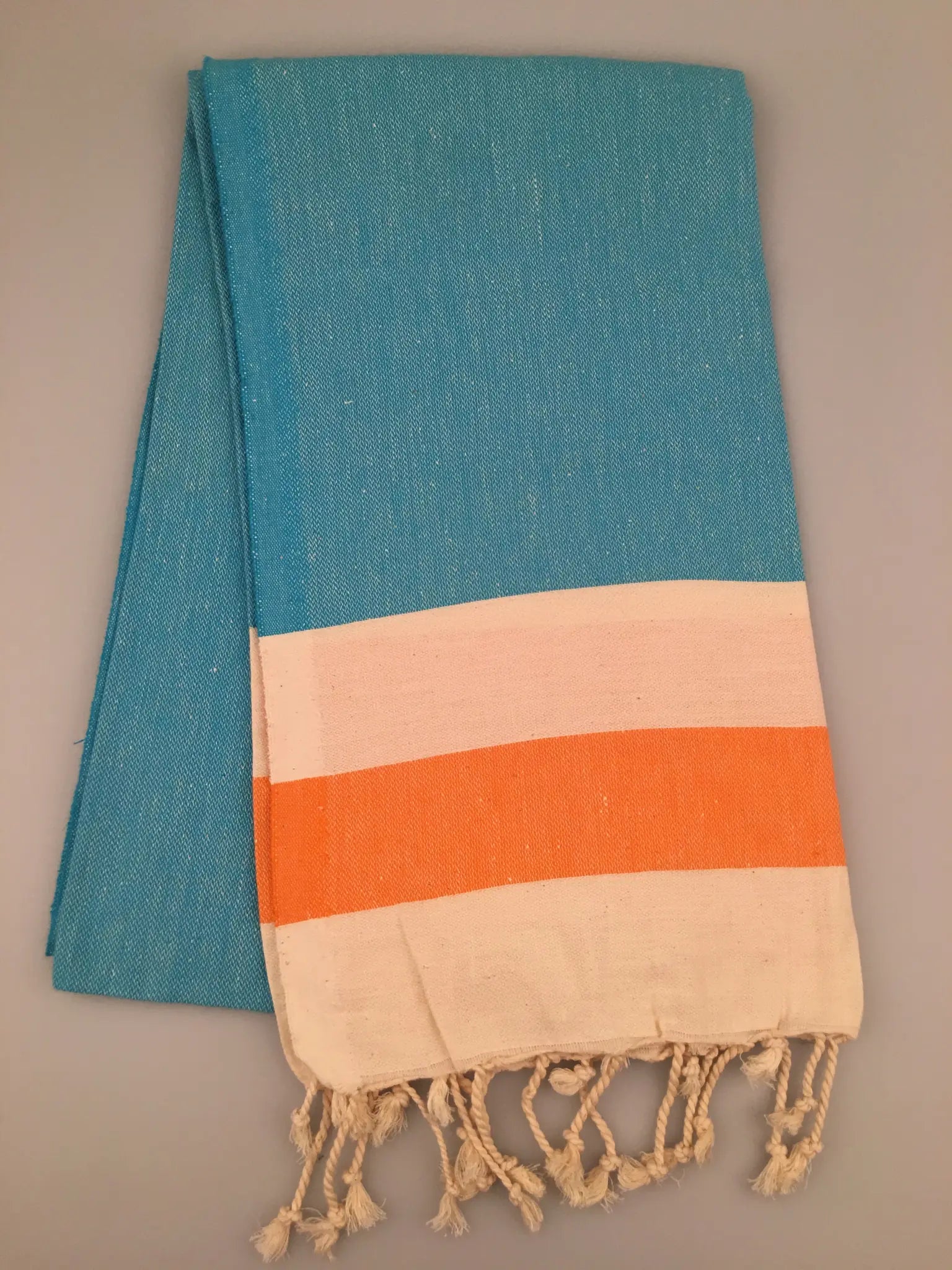 Blue Turkish towel peshtemal with orange and cream stripes, featuring fringed edges, laid flat on a neutral background.