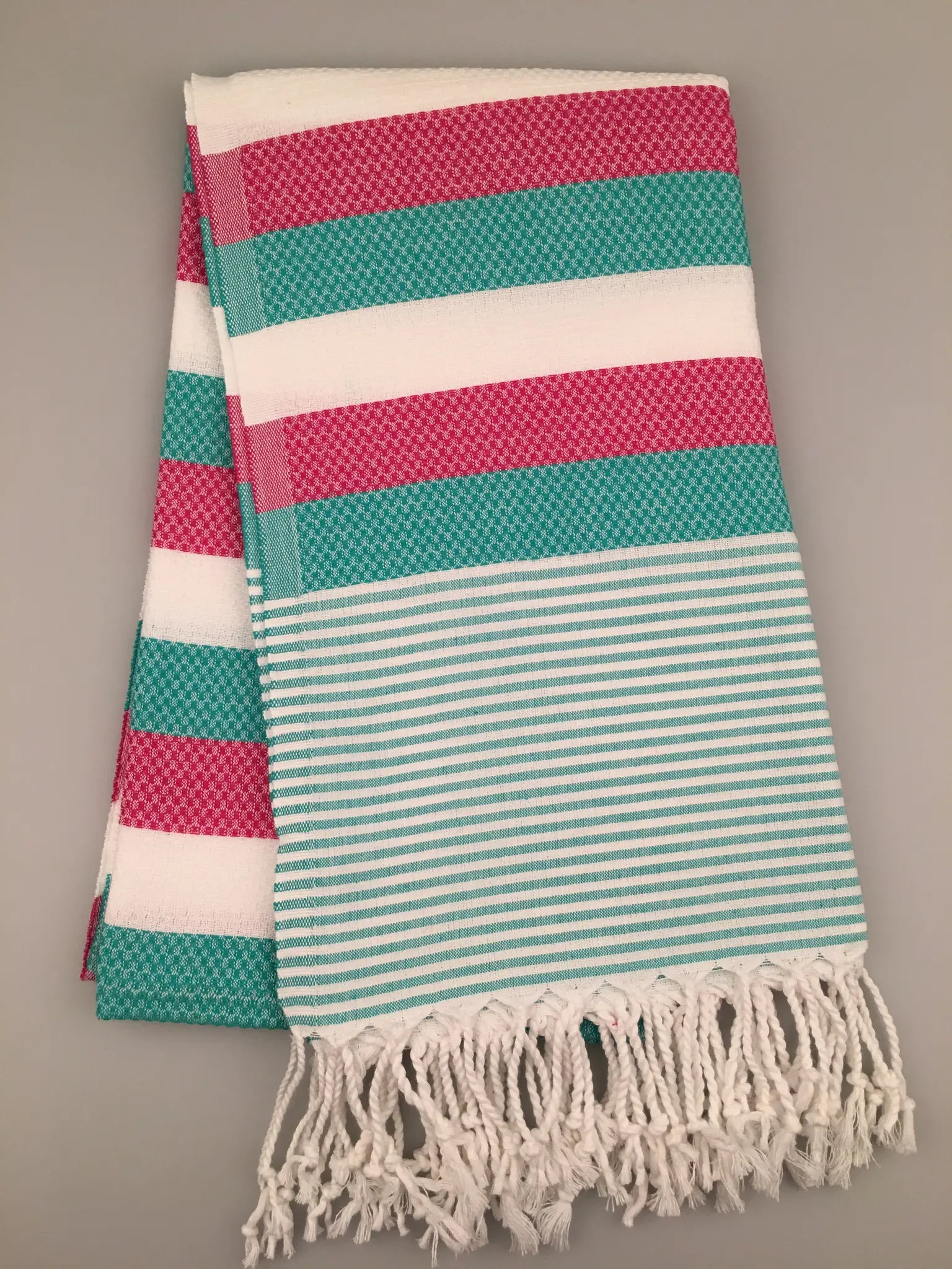 "Turquoise and pink striped Turkish towel with fringe edges, folded and laid flat on a gray background."