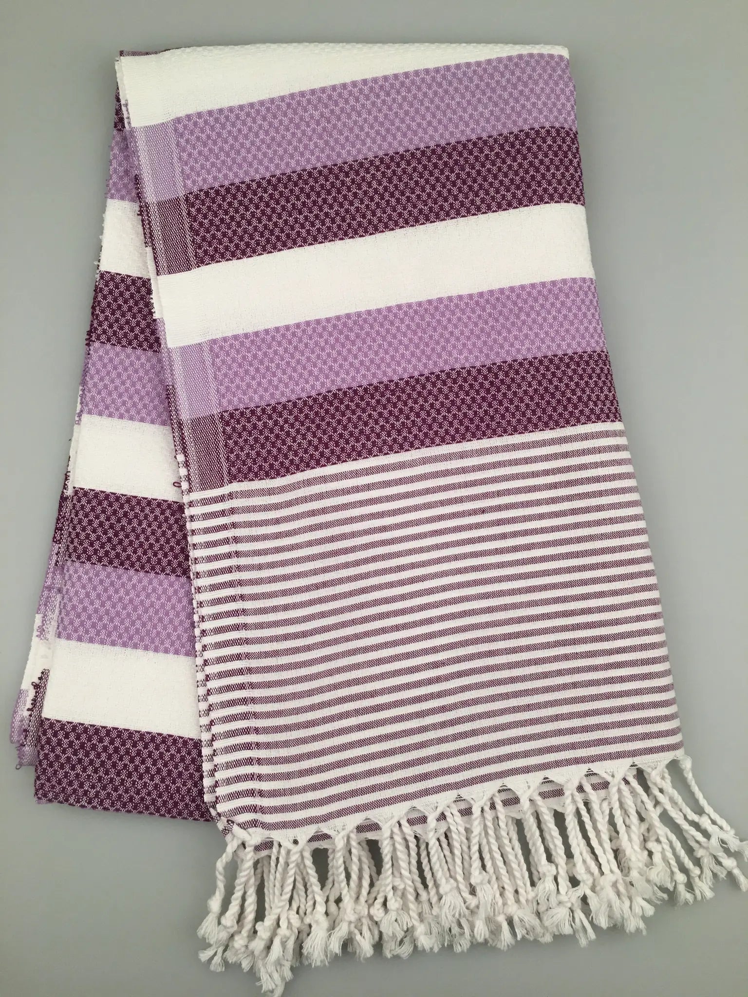 "Purple and white striped Turkish towel with textured patterns and fringed edges, laid flat on a gray background."