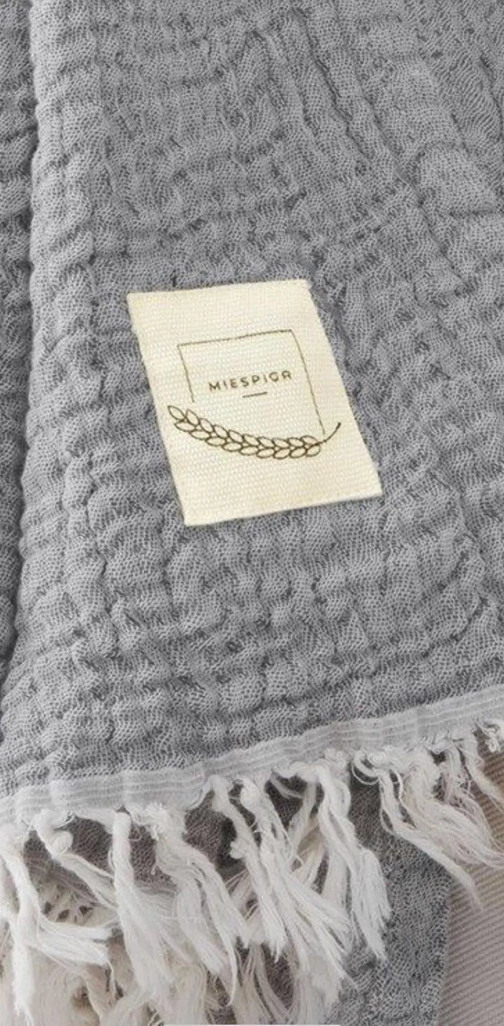 Gray textured fabric with fringed edges and a cream label featuring a gold leaf design, side view.